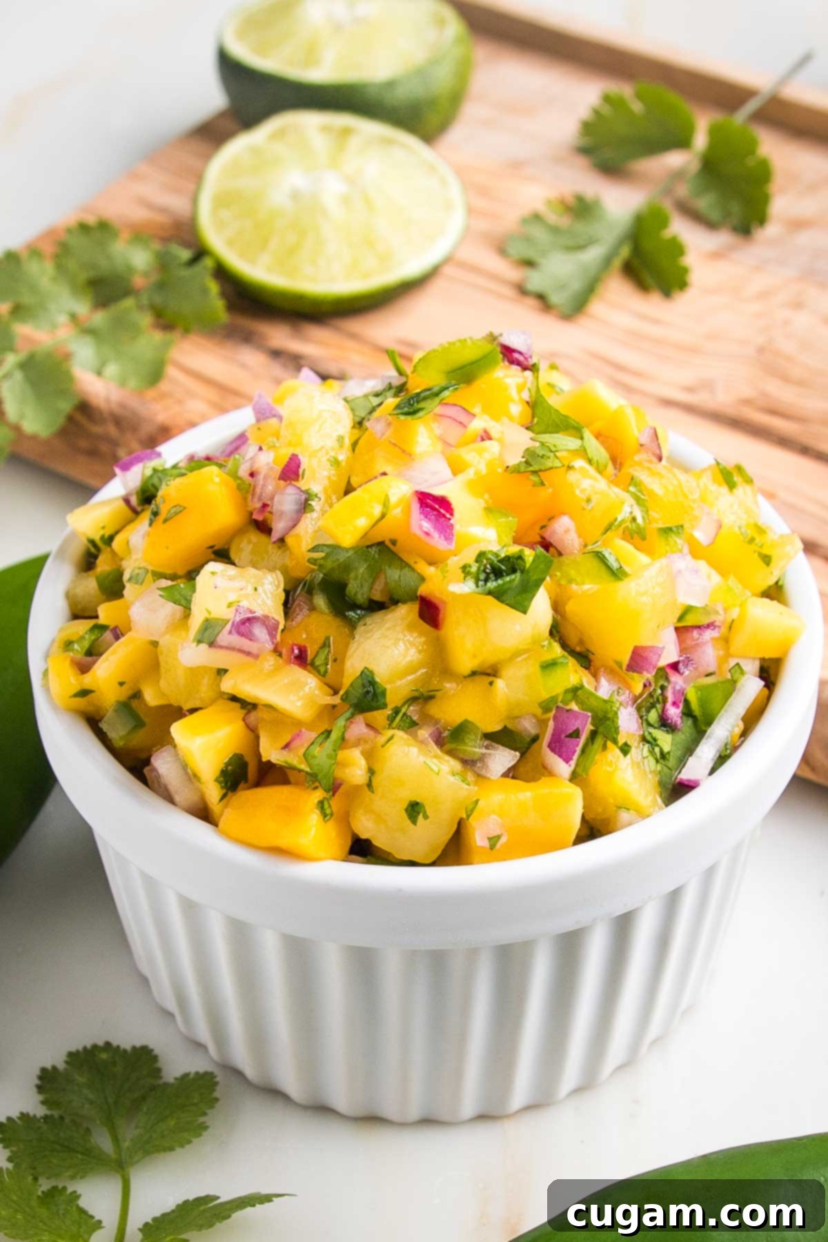 Vibrant Pineapple Mango Salsa 2 Small white bowl with pineapple mango salsa. Small pieces of bright yellow fruit, with diced red onion, jalapeno and cilantro. A juiced lime in the background with cilantro leaves on a cutting board.