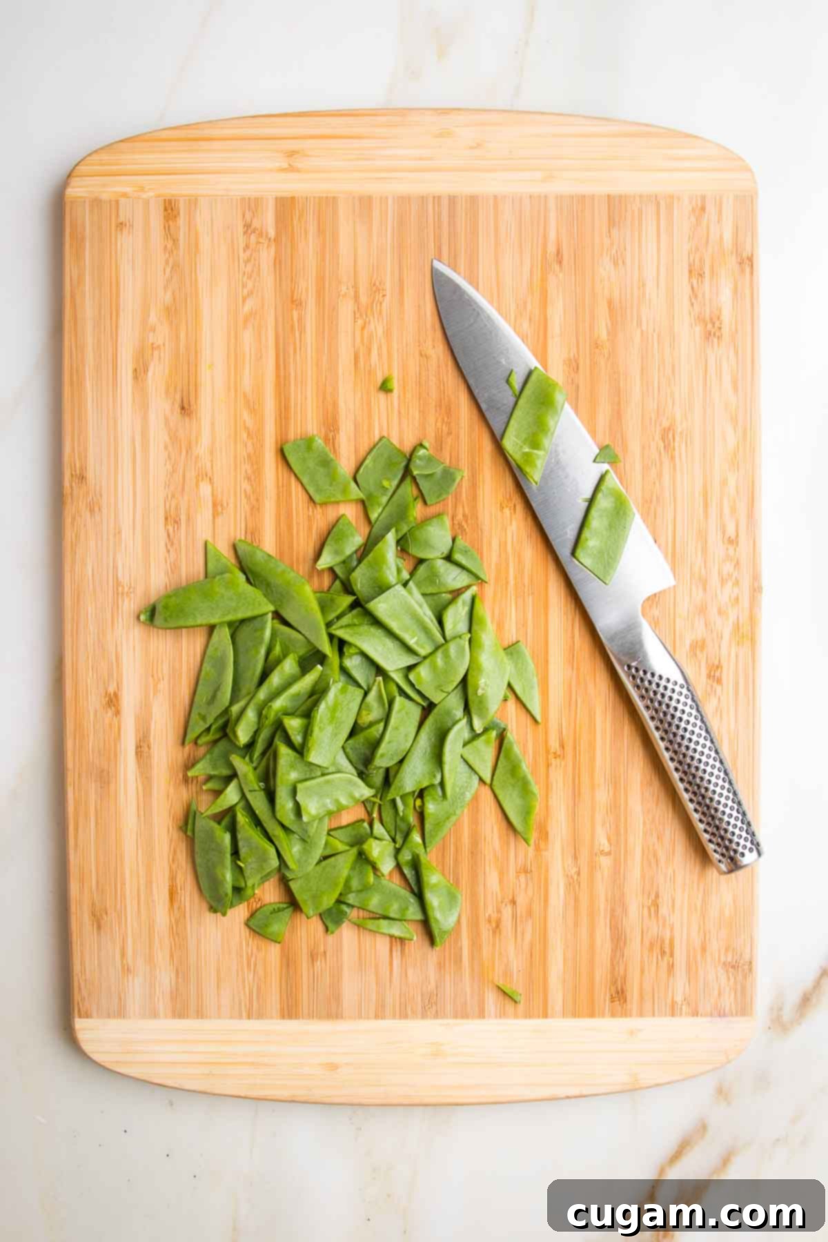 Cutting board and sharp chef's knife showing how to cut snow peas.