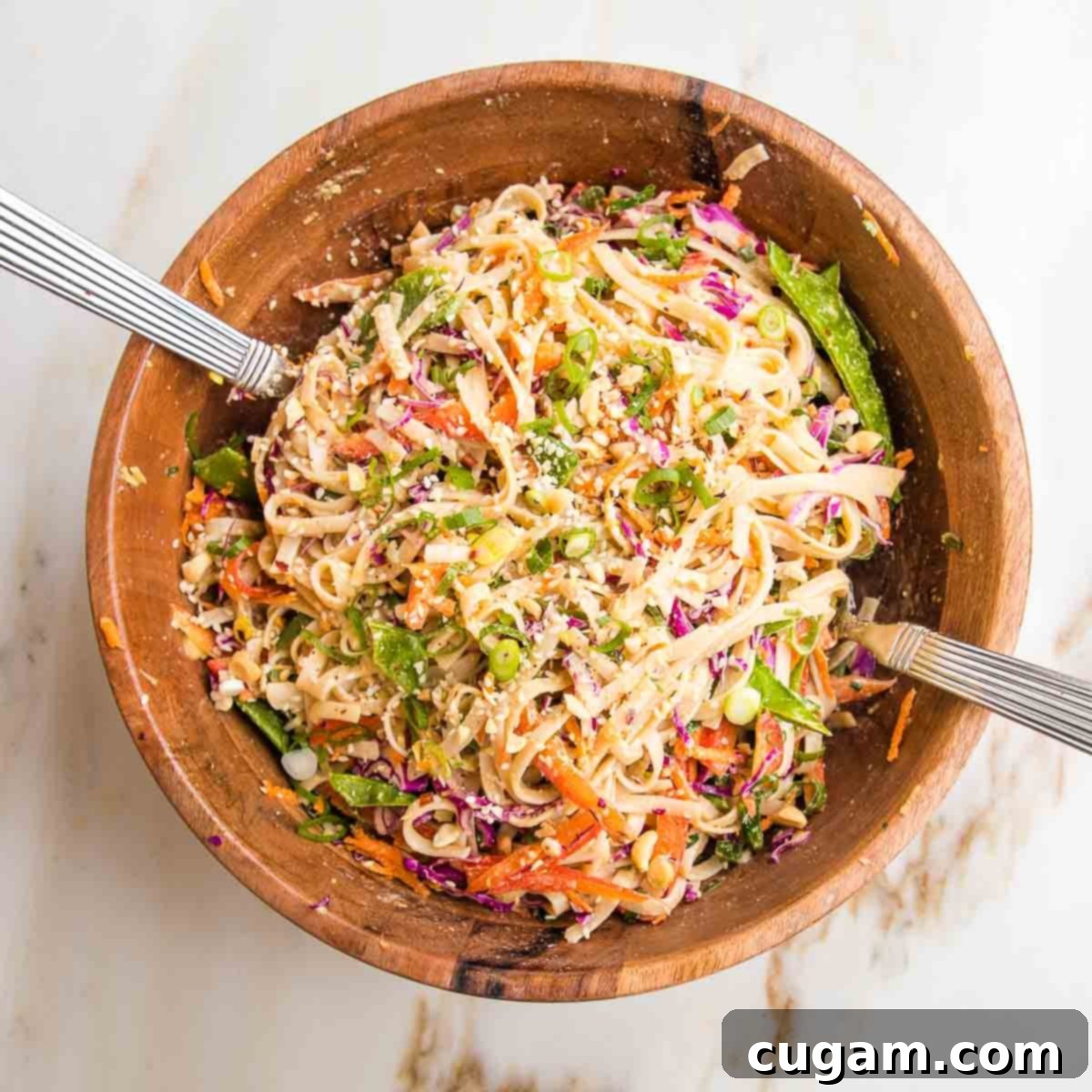 Wooden bowl filled with cold sesame noodles and brightly colored vegetables, with silver salad tossers.