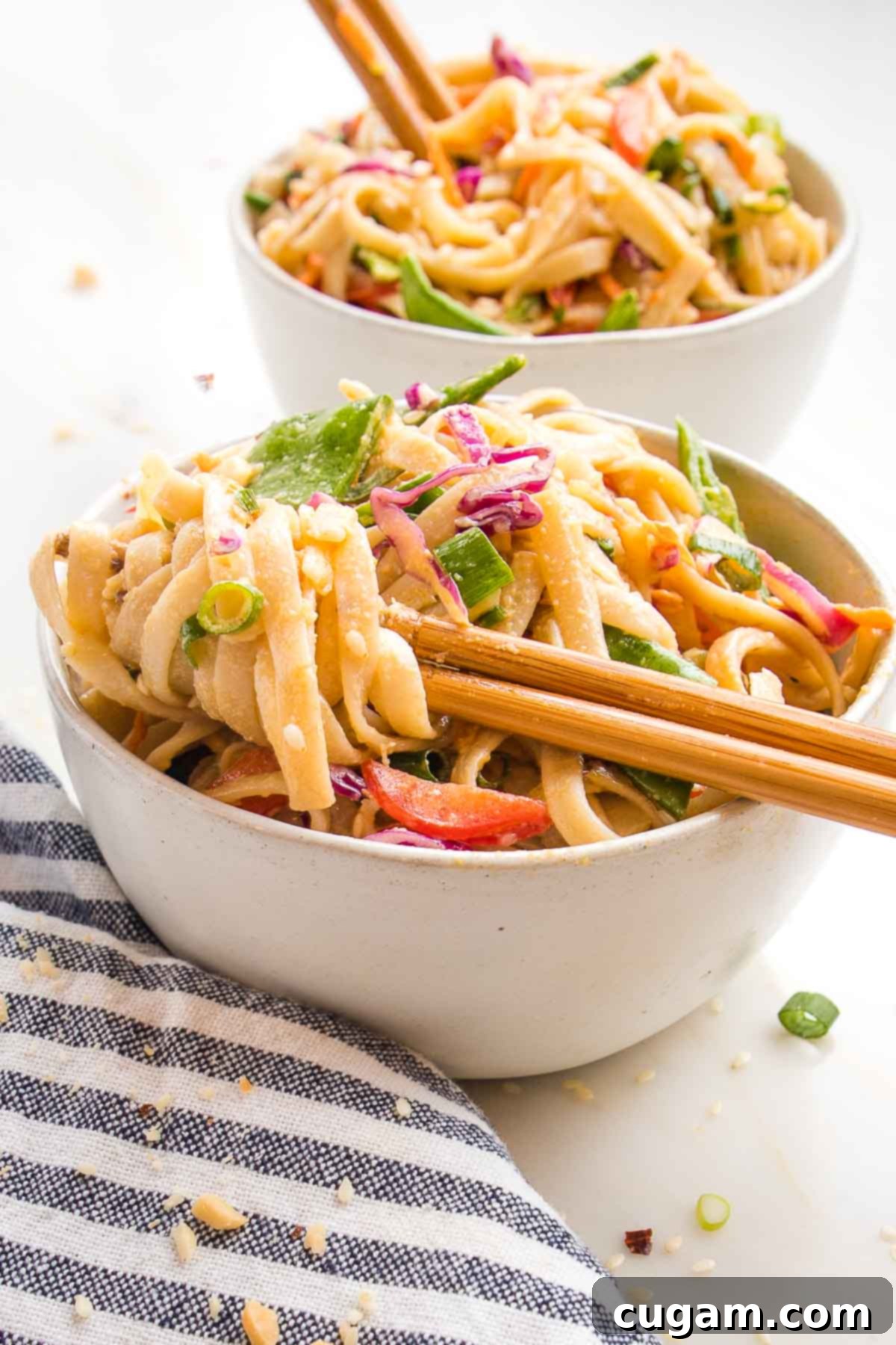 Sesame noodles twirled on a pair of chopsticks. Brightly colored chopped veggies can be seen like red peppers, shredded carrots, chopped cabbage and green onions. 