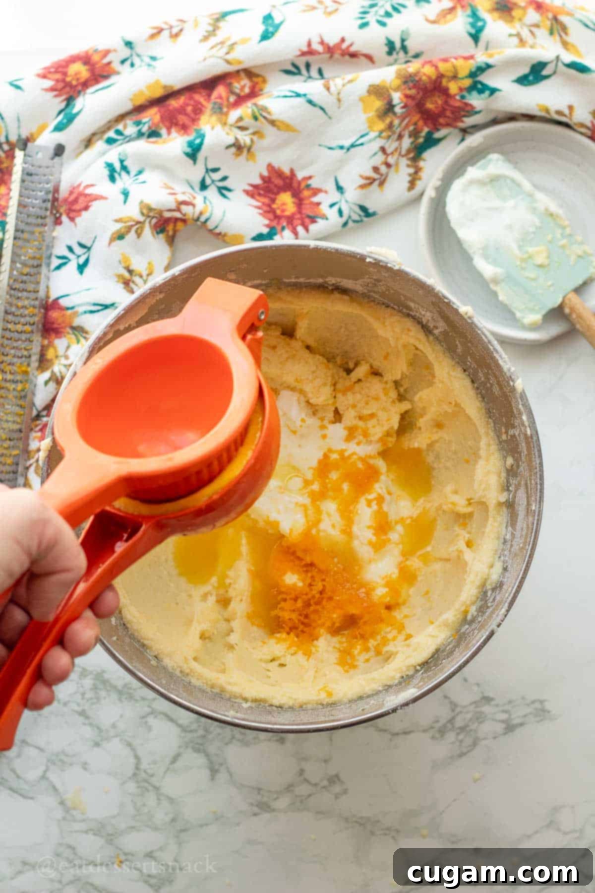 Someone squeezing fresh orange juice into a metal bowl containing orange zest and other wet ingredients.