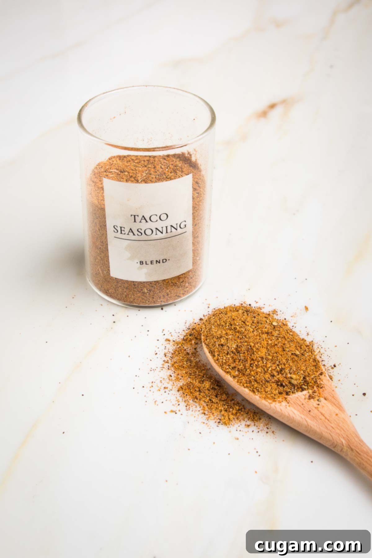 Small glass jar labeled Taco Seasoning Blend ¾ filled with orangish brown powder and a wooden spoon in front of it with same seasoning.