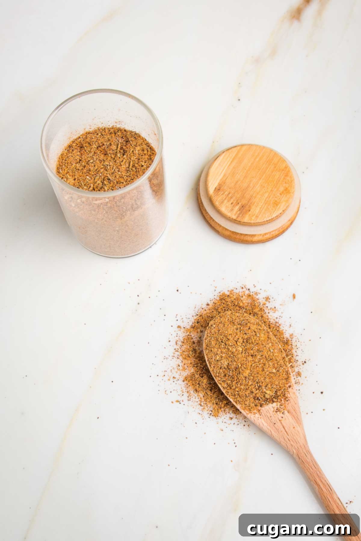 Small jar with taco seasoning blend, wooden top off to the side. Wooden spoon filled with seasoning and some around the spoon on the counter.