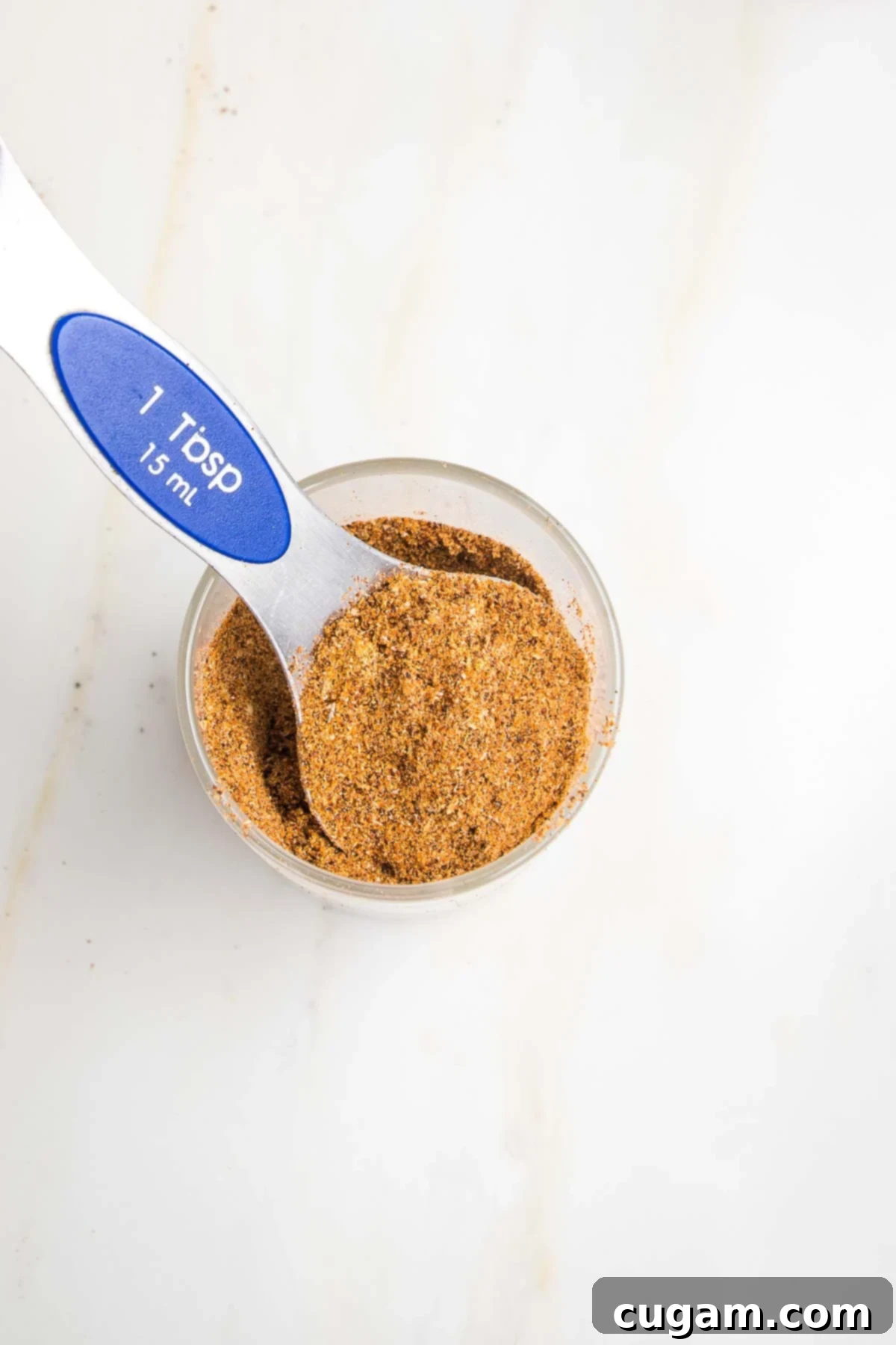 Small glass jar of taco seasoning with a 1 Tablespoon measuring spoon scooping some out of the jar. 
