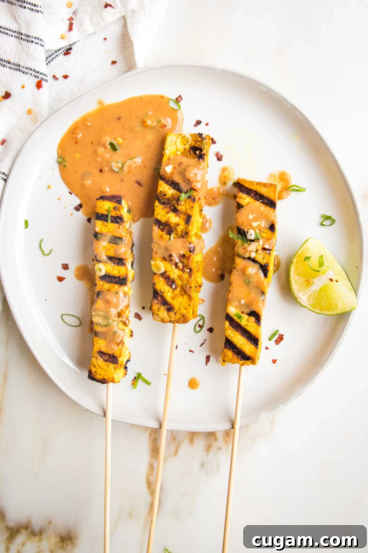 Large white plate with three tofu satay skewers. Tofu has grill marks and peanut sauce is drizzled onto them. There is a lime wedge and a puddle of additional sauce. 