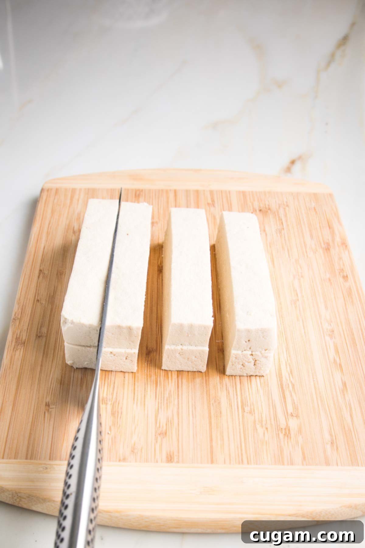 Drained tofu on a wooden board, slicing through to produce 8 equal long satay slices.