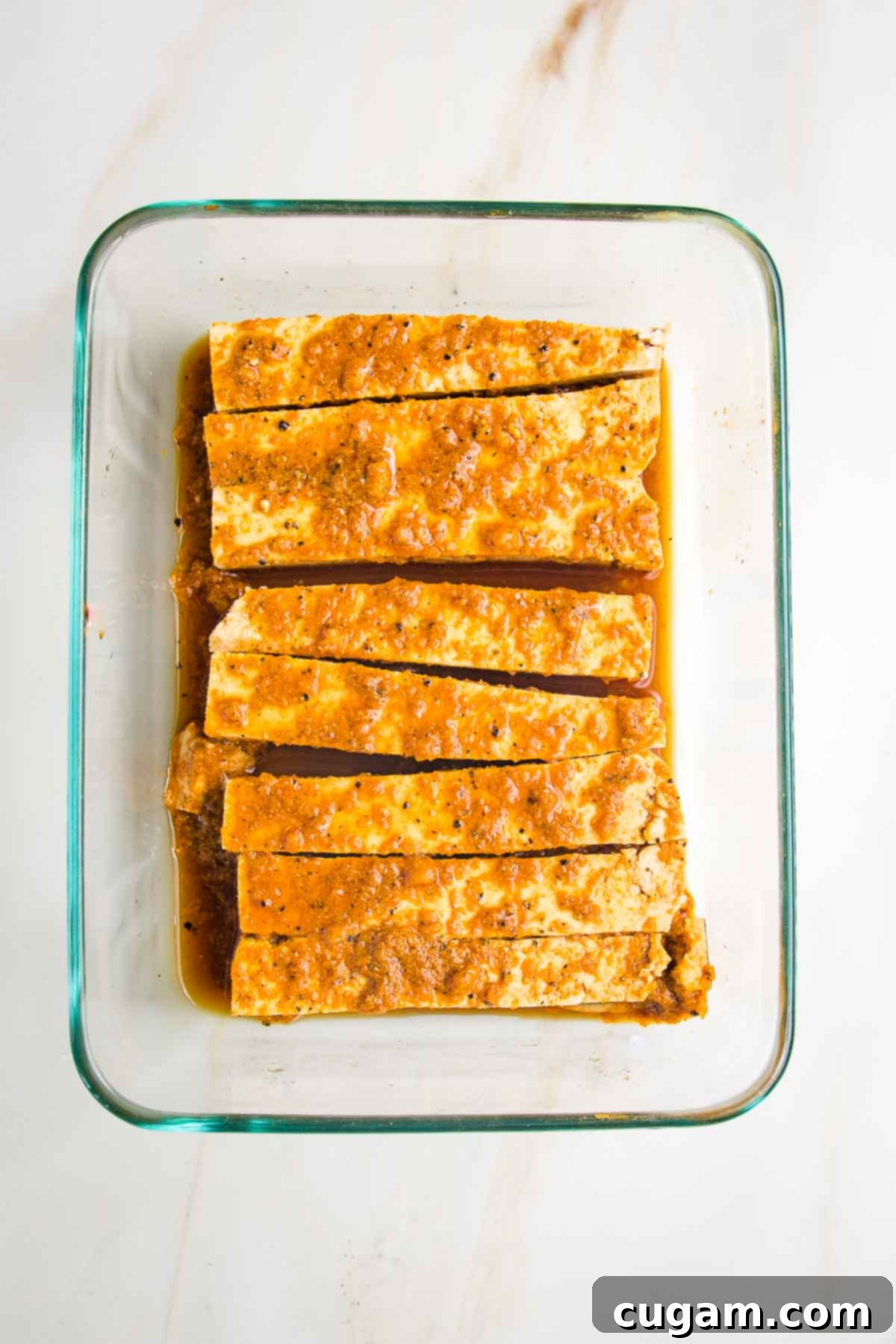 Long thin slices of tofu for satay with brownish-orange marinade in a rectangular glass dish.