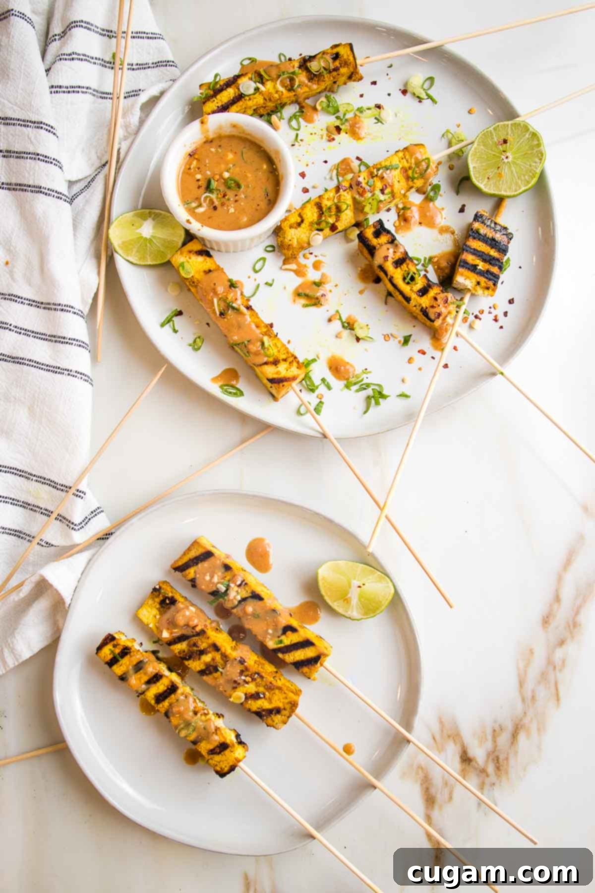 Plate filled with tofu satay skewers, peanut sauce and lime wedges. Everything is sprinkled with chili flakes, sliced scallions and crushed peanuts.  A smaller plate with three skewers and a lime wedge is on the bottom. 