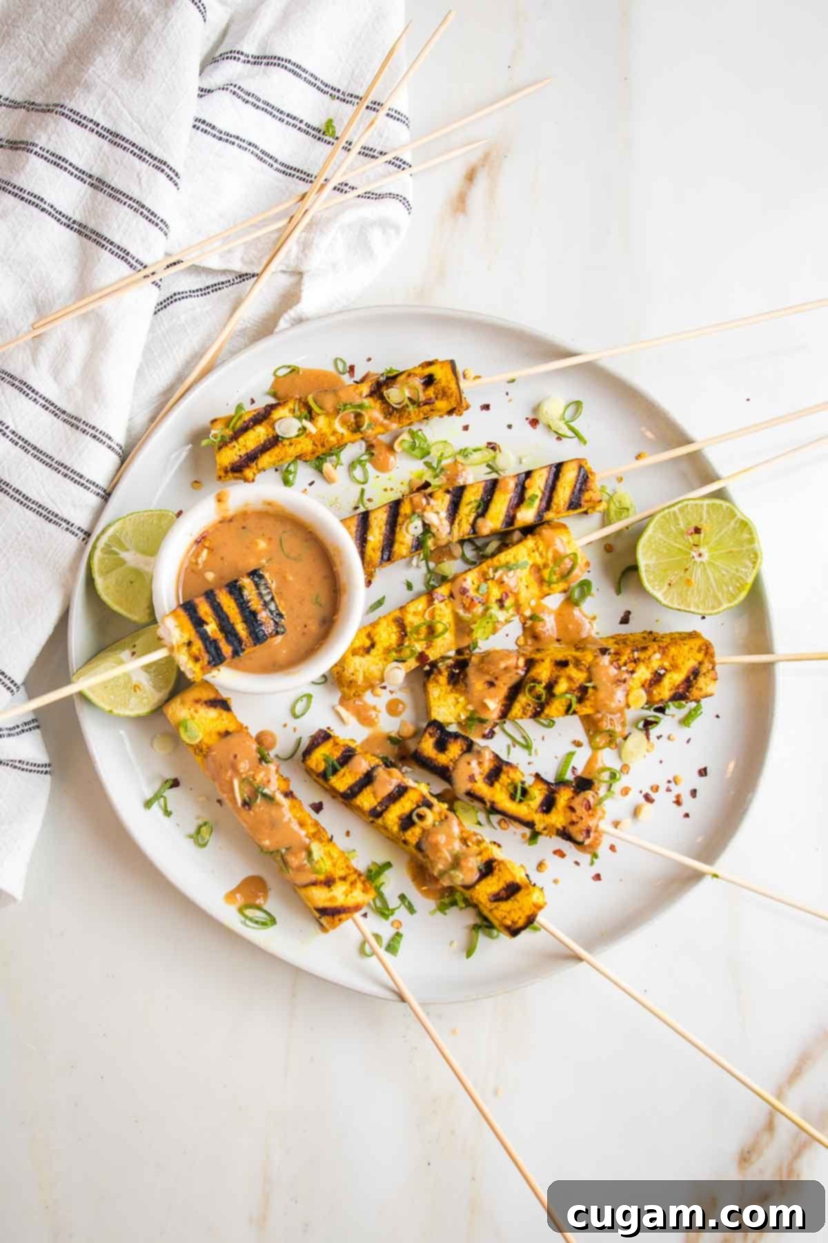 Plate of grilled tofu satay skewers with a small dish of peanut sauce, sauce drizzled onto skewers and everything is sprinkled with scallions, peanuts, crushed red pepper and more sauce. 