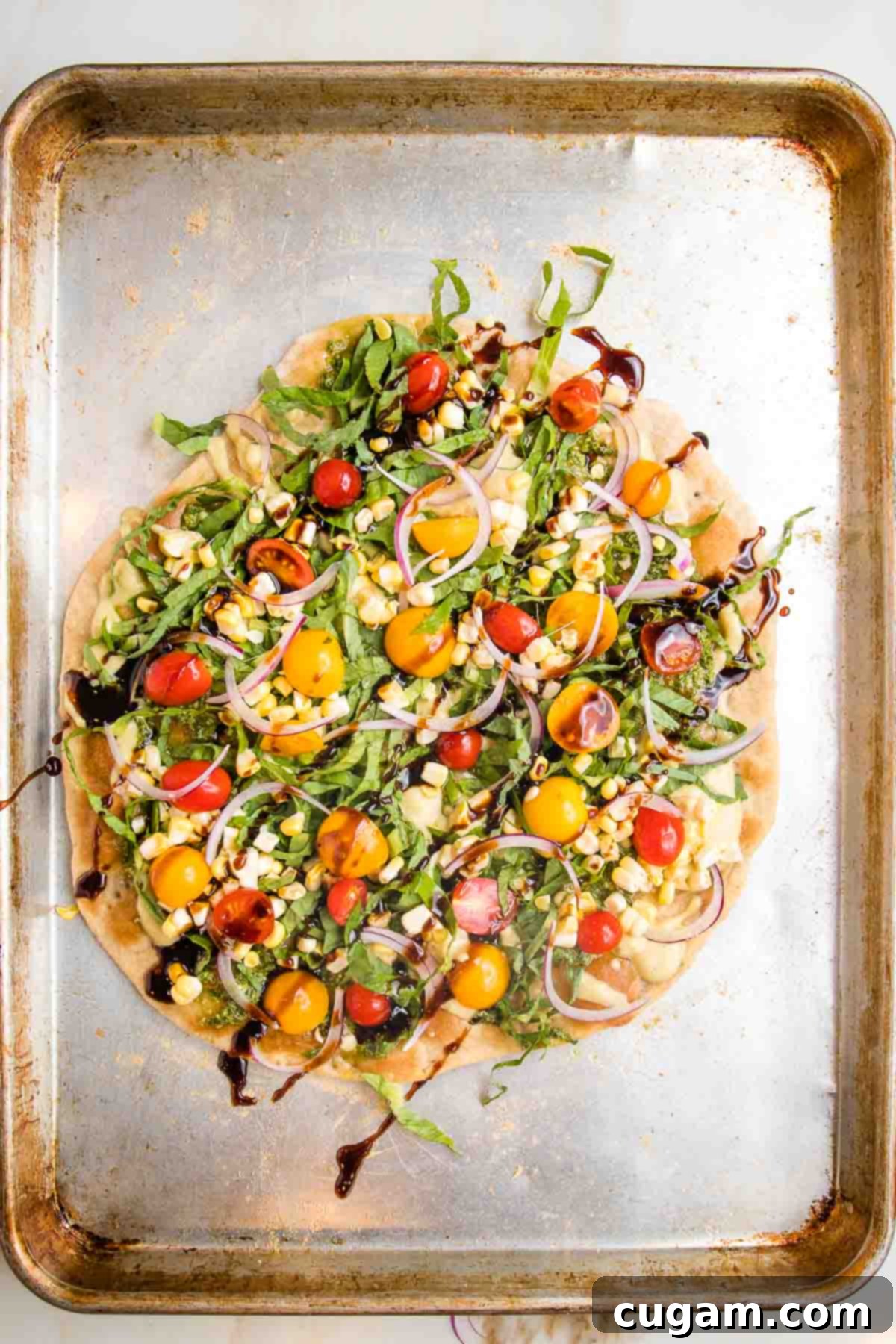 Flatbread pizza on a baking tray. Toppings include pesto, hummus, red and yellow tomatoes, chopped greens and a balsamic drizzle.
