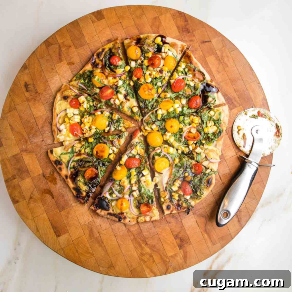 Flatbread pizza topped with red and yellow tomatoes, greens, onions and pesto. Pizza cutter on the side of cutting board holding sliced flatbread.