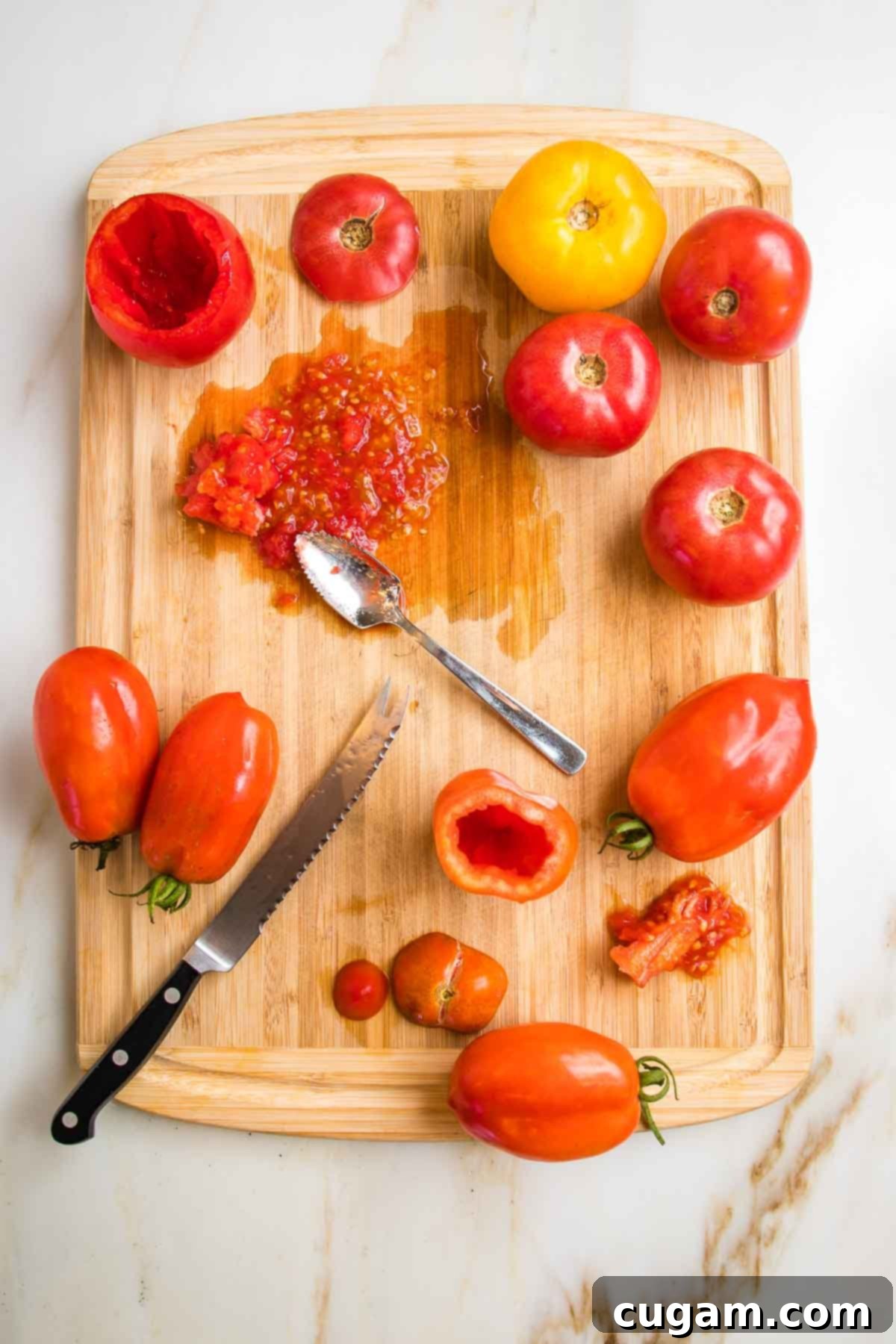 Wooden cutting board with tomatoes, tomato knife and grapefruit spoon. Some tomatoes have their tops cut off and seeds scooped out.