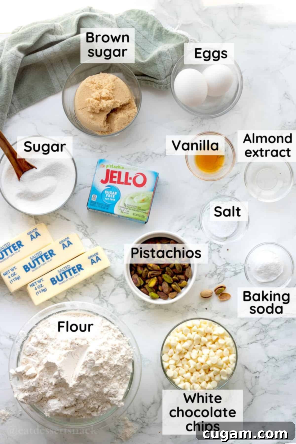 Various ingredients for pistachio pudding cookies, including butter, sugars, pistachios, and pudding mix, displayed in glass bowls on a marble countertop.