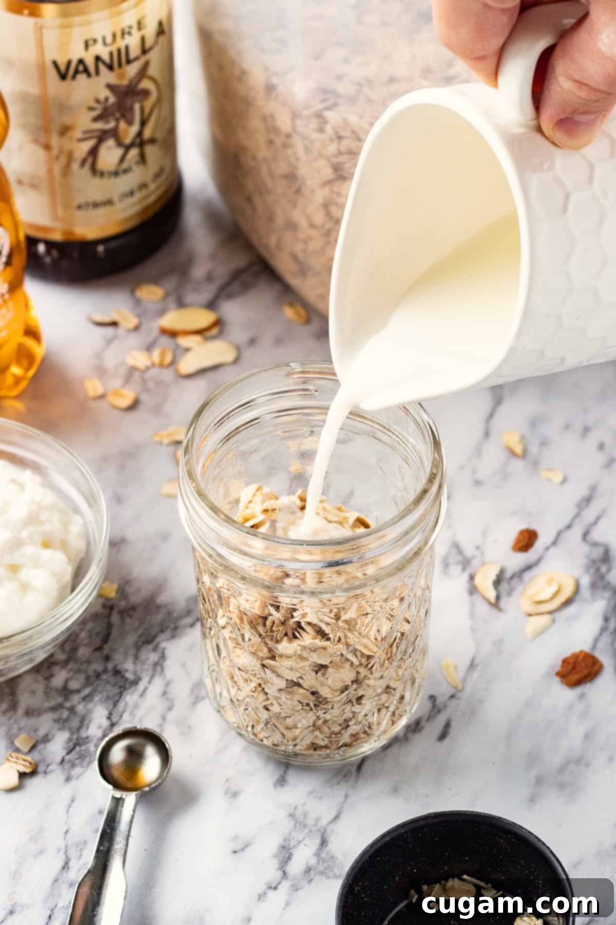 Pouring milk into jar of oats with vanilla dn yogurt nearby on marble countertop.