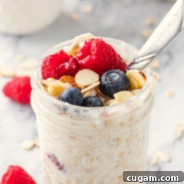 Jar of vanilla overnight oats topped almonds, rapsberries and blueberries with metal spoon.