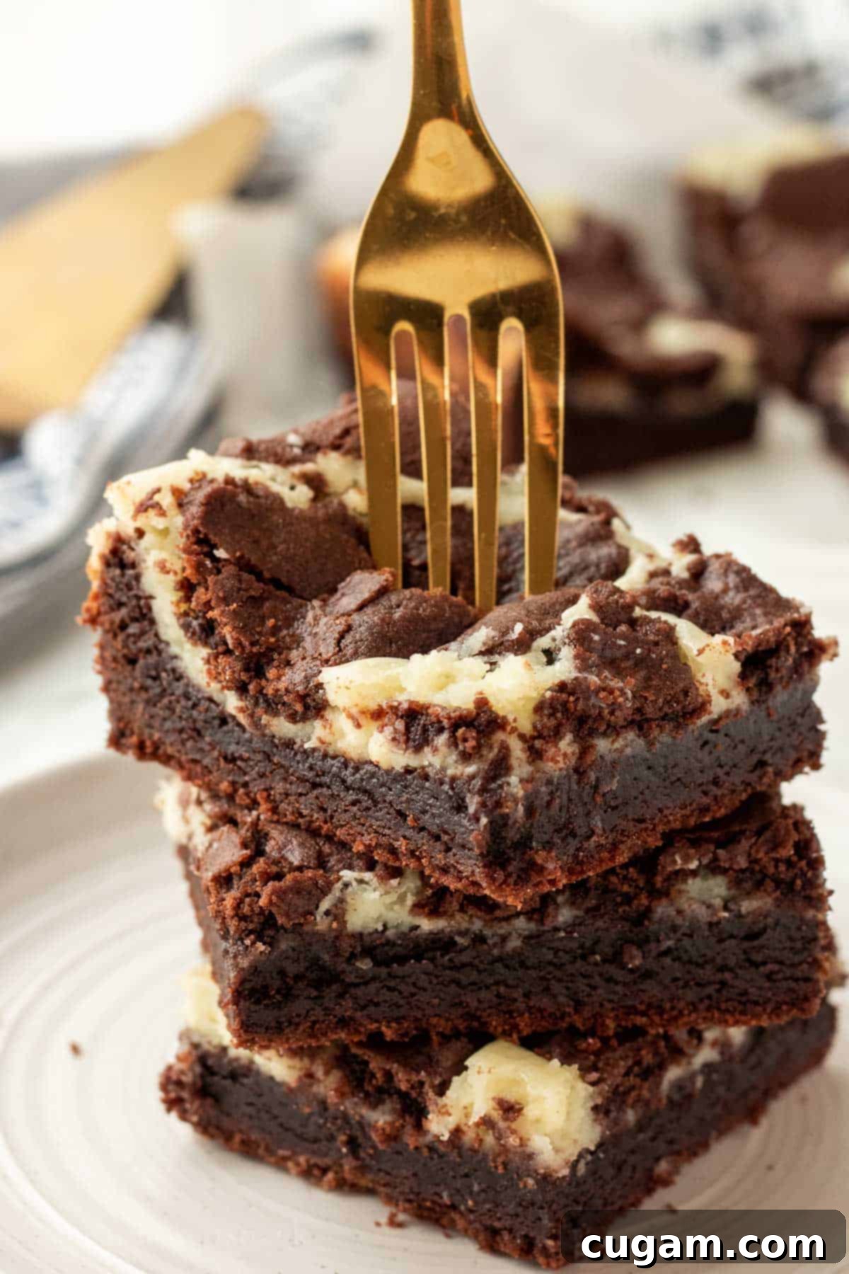 Stack of 3 baked cheesecake brownie bars with gold fork, showcasing the layers of fudgy brownie, creamy cheesecake, and soft brownie crumble.