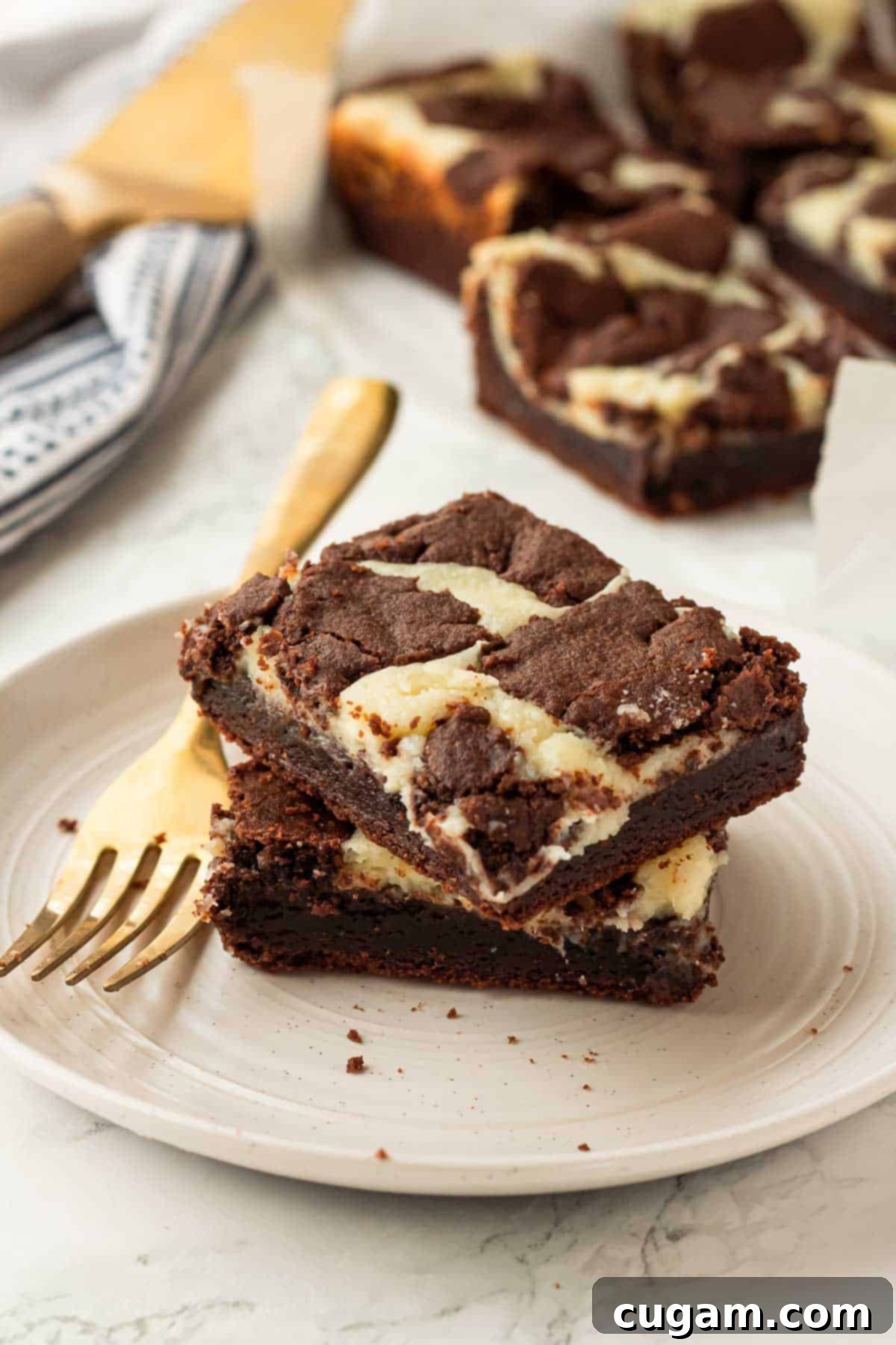 Stack of 2 baked cheesecake brownie bars on a white plate with a gold fork lying near them, highlighting their rich layers.