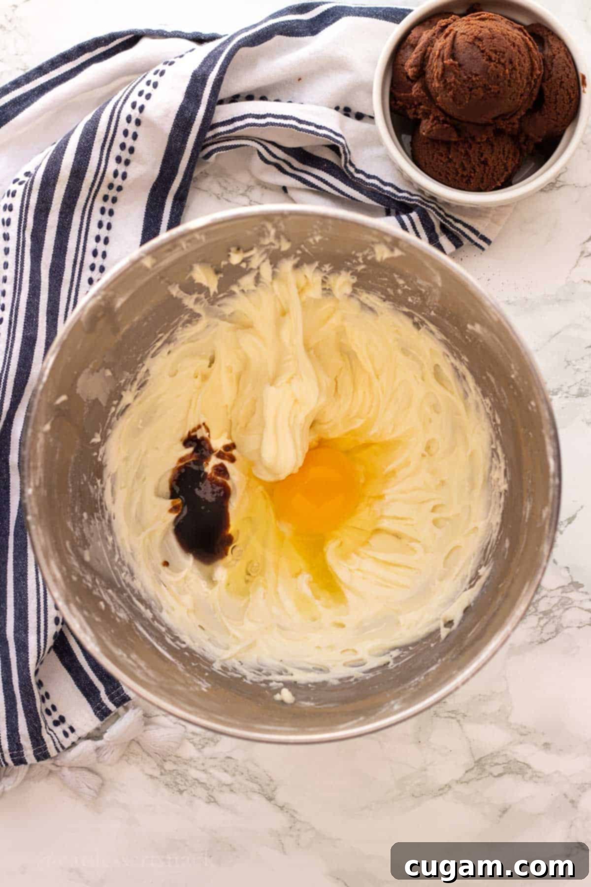 Whipped cream cheese and powdered sugar in a metal bowl, with an egg and vanilla extract on top, ready for mixing to create the cheesecake filling.