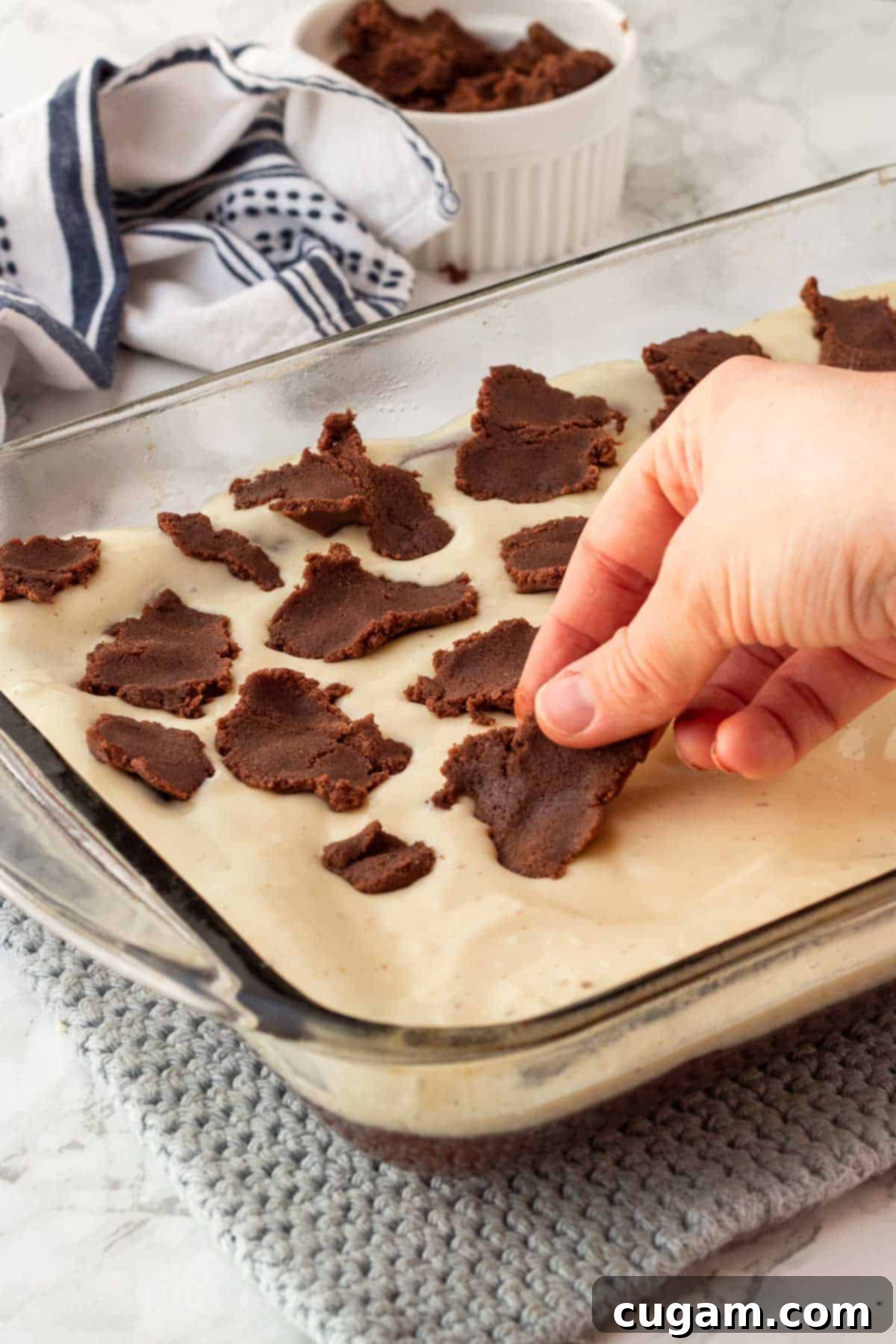 A hand gently placing small, flattened pieces of raw brownie dough on top of the smooth cream cheese filling, creating a decorative layer.
