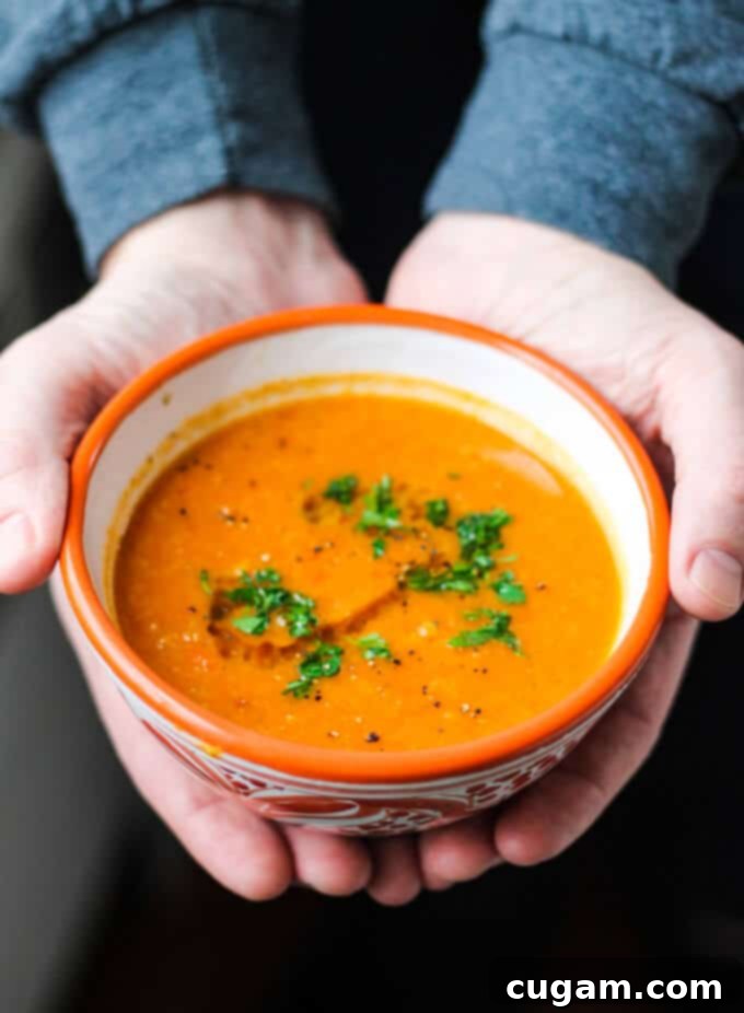 Hands holding Tomatoes Red Lentils Coconut Soup in orange bowl, garnished with fresh herbs
