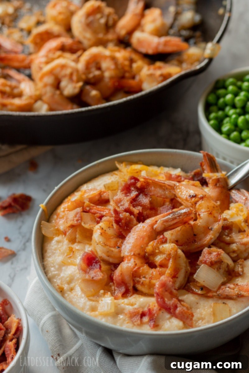 Gray bowl with cheesy grits, shrimp, and bacon, with a cast iron pan filled with shrimp and onions in the background.