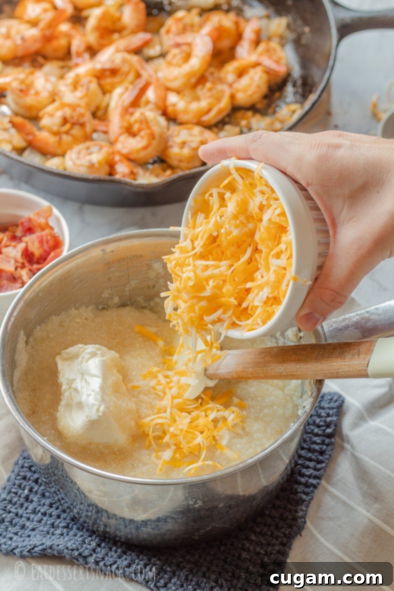 Hand pouring shredded cheddar cheese into a pan of cooked grits and cream cheese, creating a rich, gooey texture.