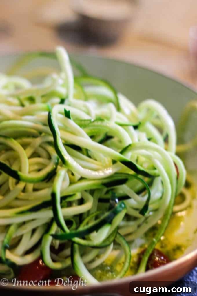 Freshly spiralized zucchini noodles being added to a pan with cherry tomatoes and pesto sauce.