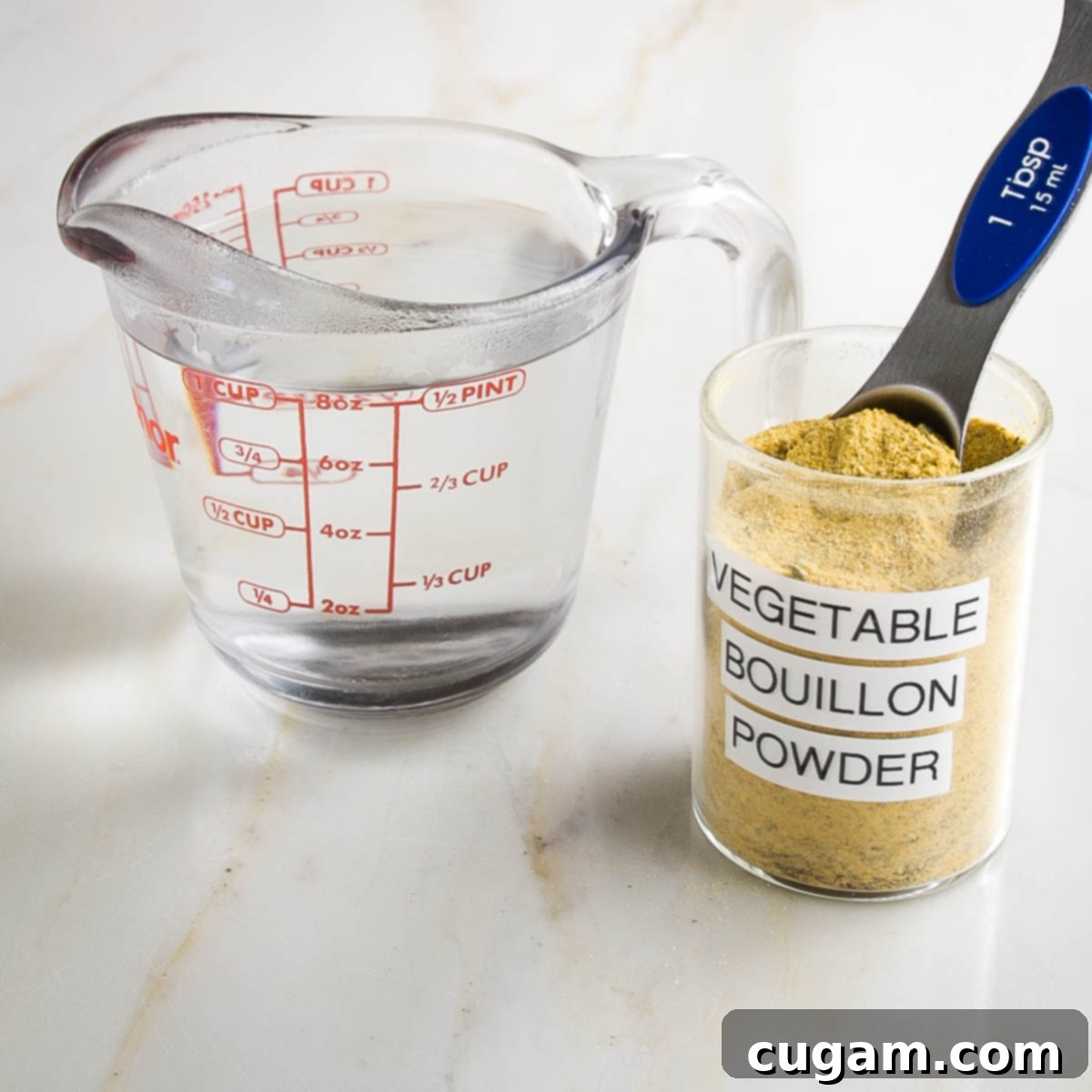 Glass measuring cup with 1 cup water. Spice jar labeled "vegetable bouillon powder" with Tablespoon measuring spoon in it.