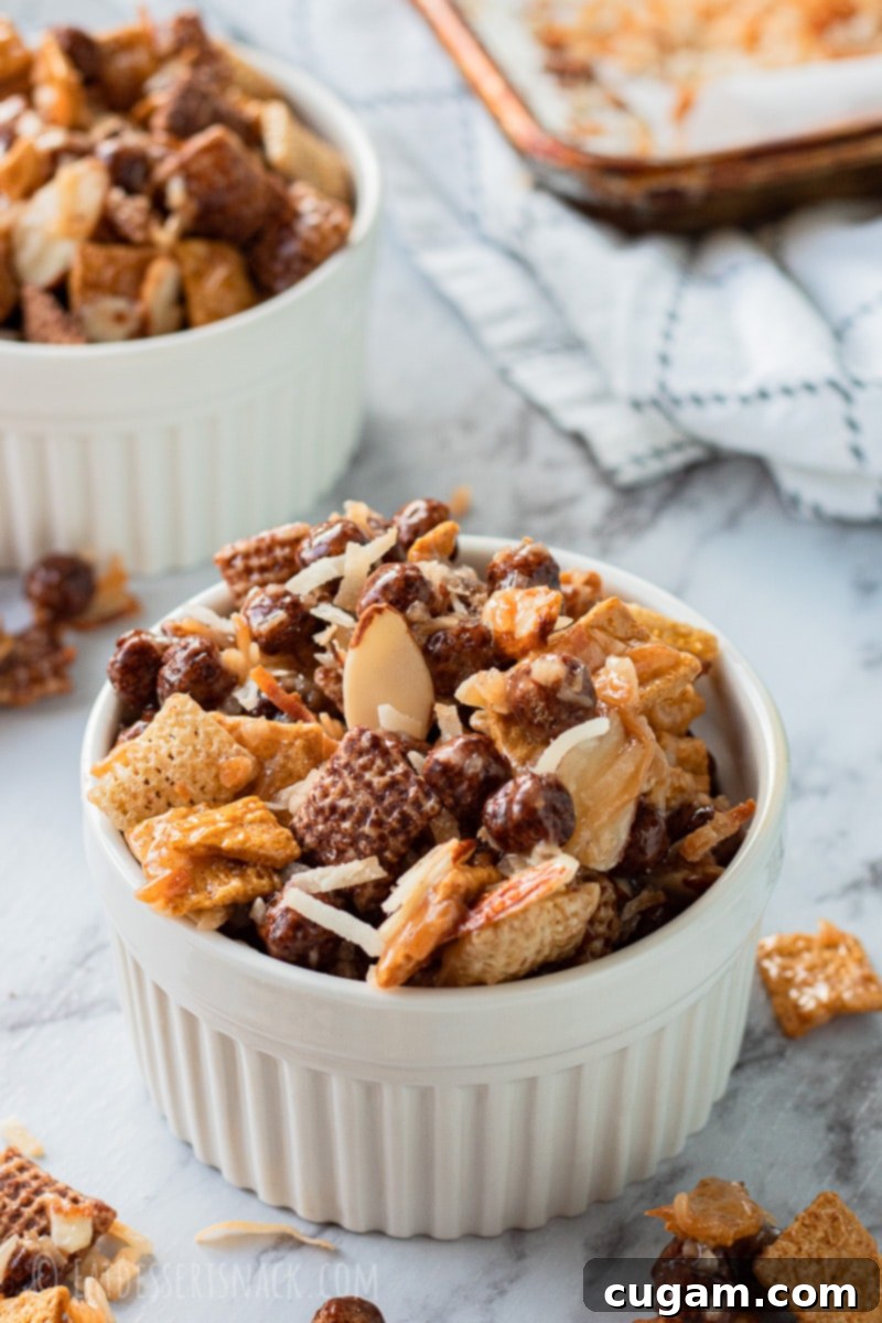 A large white bowl filled with glistening Chocolate Gooey Chex Mix, featuring visible pieces of chocolate cereal, toasted coconut, and almonds, presented beautifully on a clean surface.