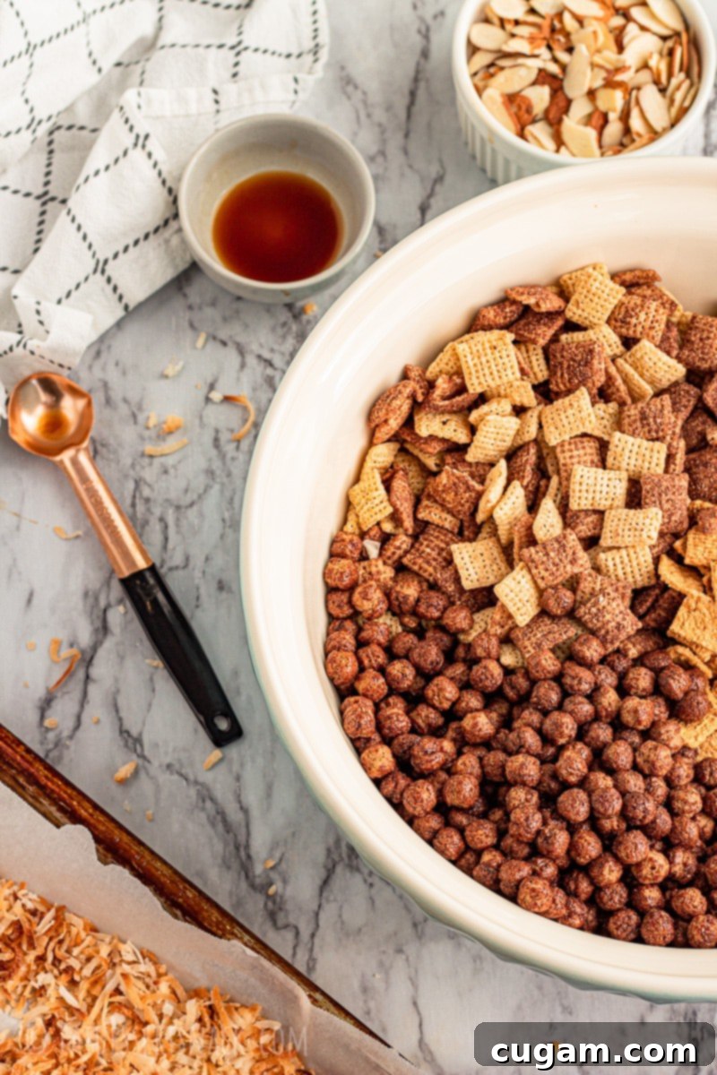 A collection of ingredients for Chocolate Gooey Chex Mix, neatly arranged: toasted coconut in a pan, a small bowl of vanilla extract, a copper measuring spoon, a large bowl containing Cocoa Puffs and Chocolate Chex, and a white bowl of slivered almonds.