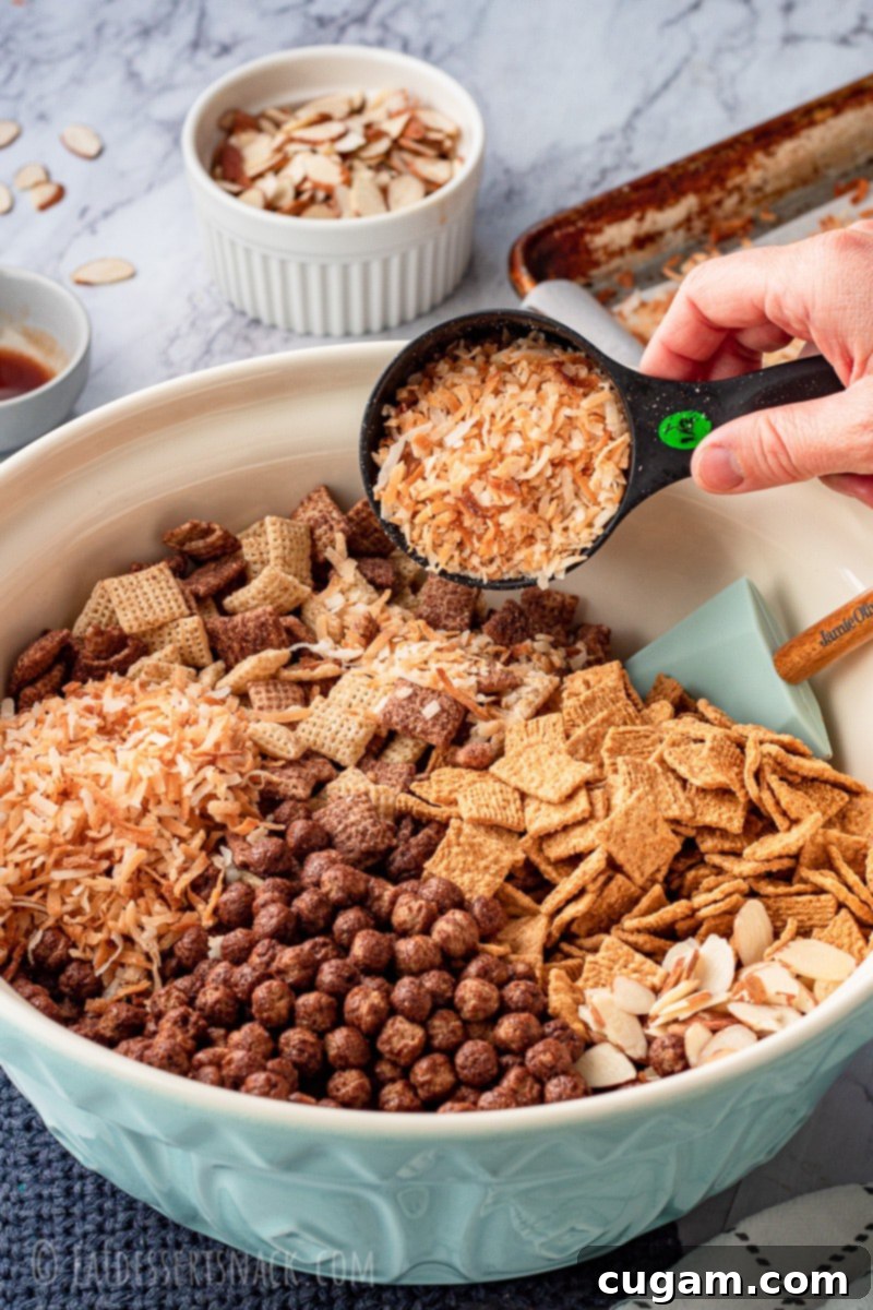 A clear measuring cup carefully pouring golden toasted coconut into a large blue bowl filled with a mixture of different cereals, indicating the start of the Chex mix preparation.
