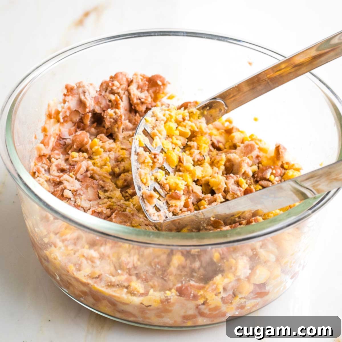 Using a potato masher to mash beans in a glass mixing bowl, creating a chunky texture.