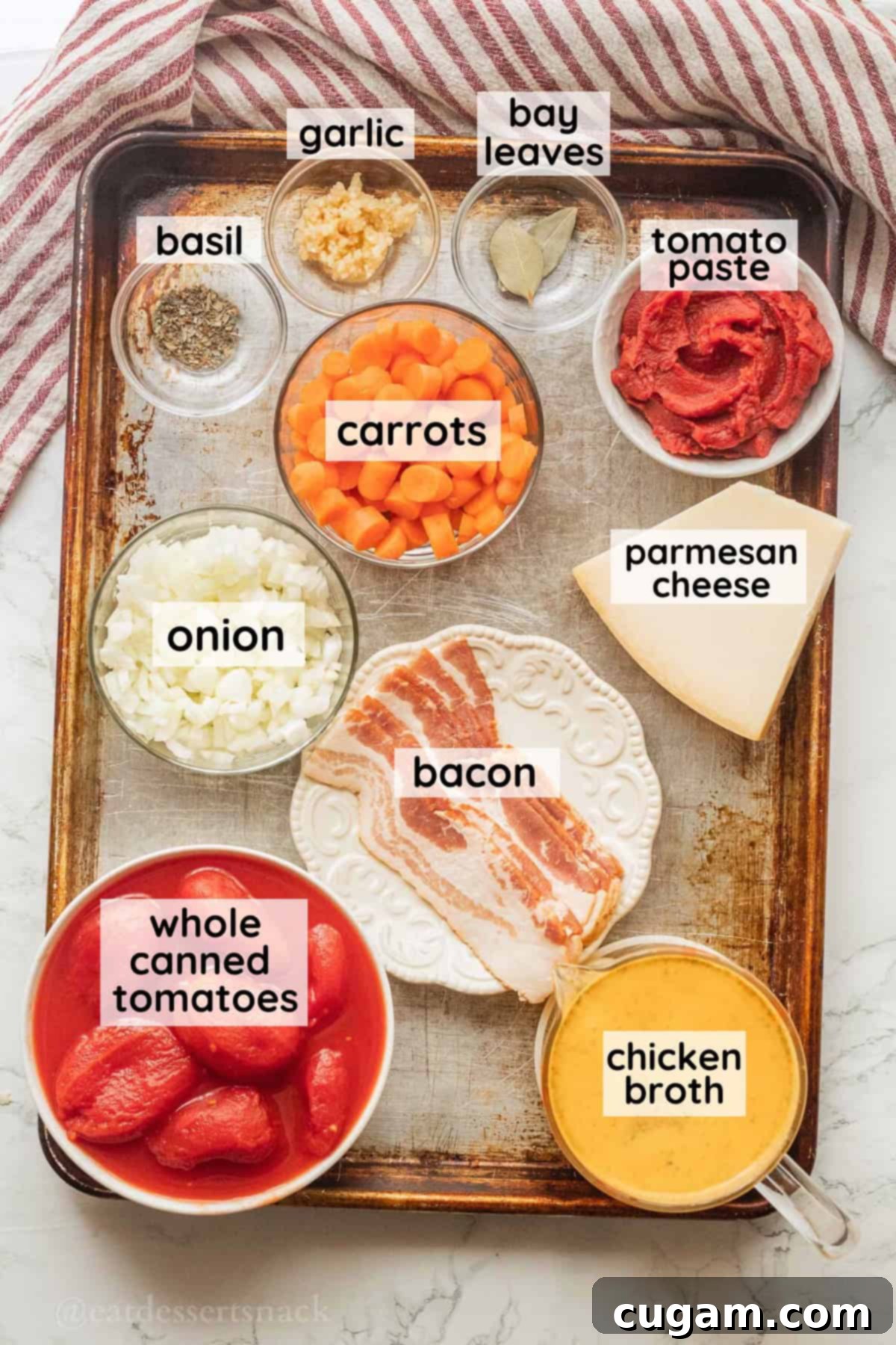 Various ingredients for canned tomato soup, including bacon, carrots, onions, tomato paste, canned whole peeled tomatoes, and herbs, arranged in glass bowls and jars on a wooden surface, clearly labeled.