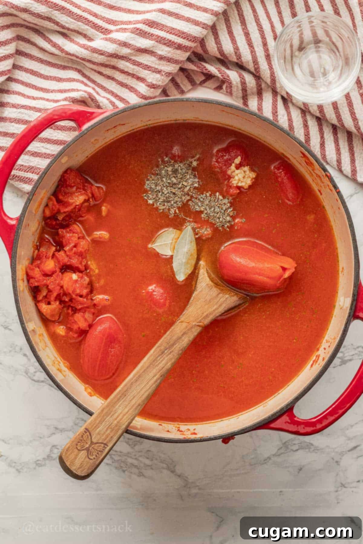 A red pot filled with tomato soup ingredients, including whole peeled tomatoes, chicken broth, bay leaves, minced garlic, and dried basil, before simmering.