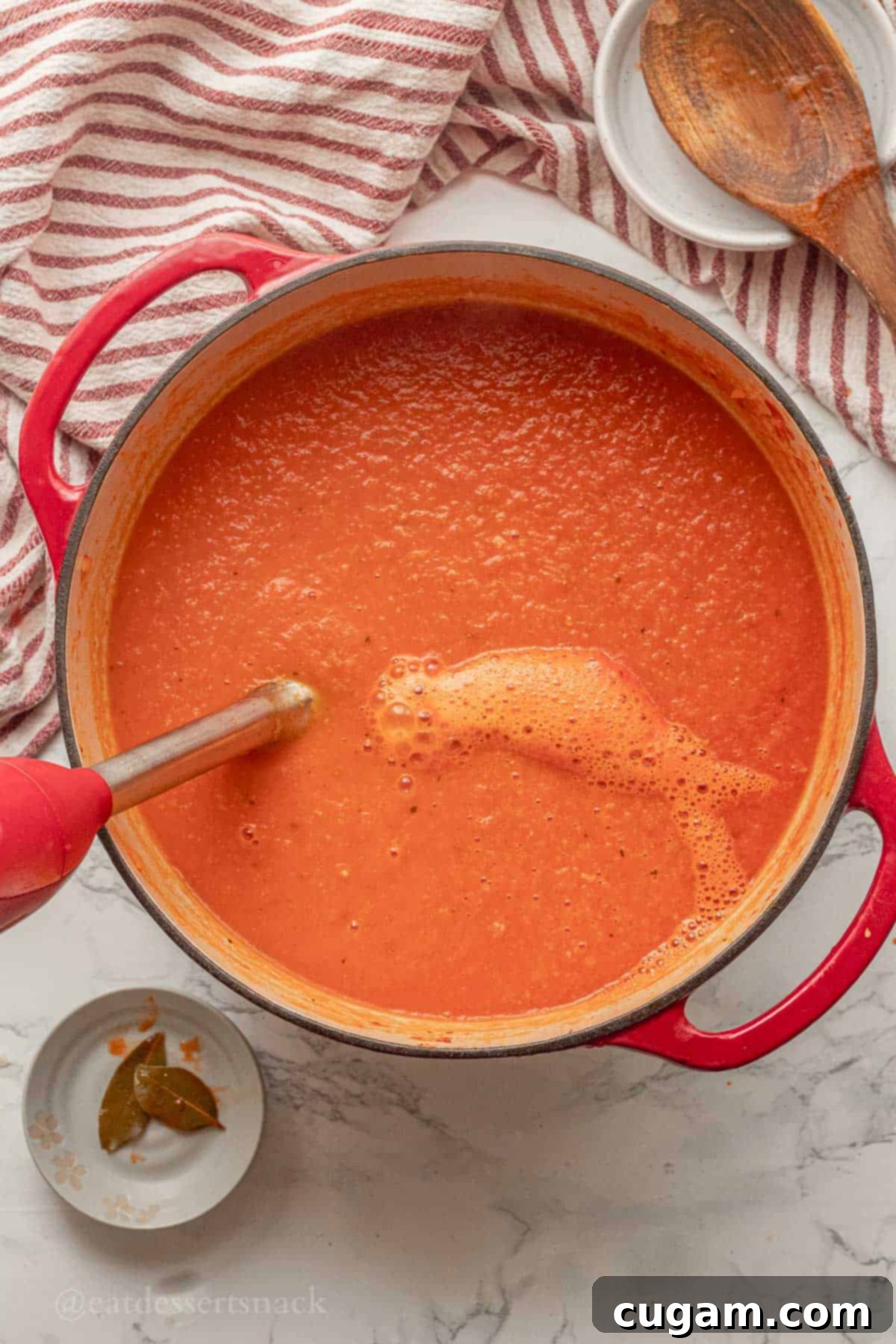 An immersion blender actively blending cooked tomato soup in a red pot, creating a smooth consistency.