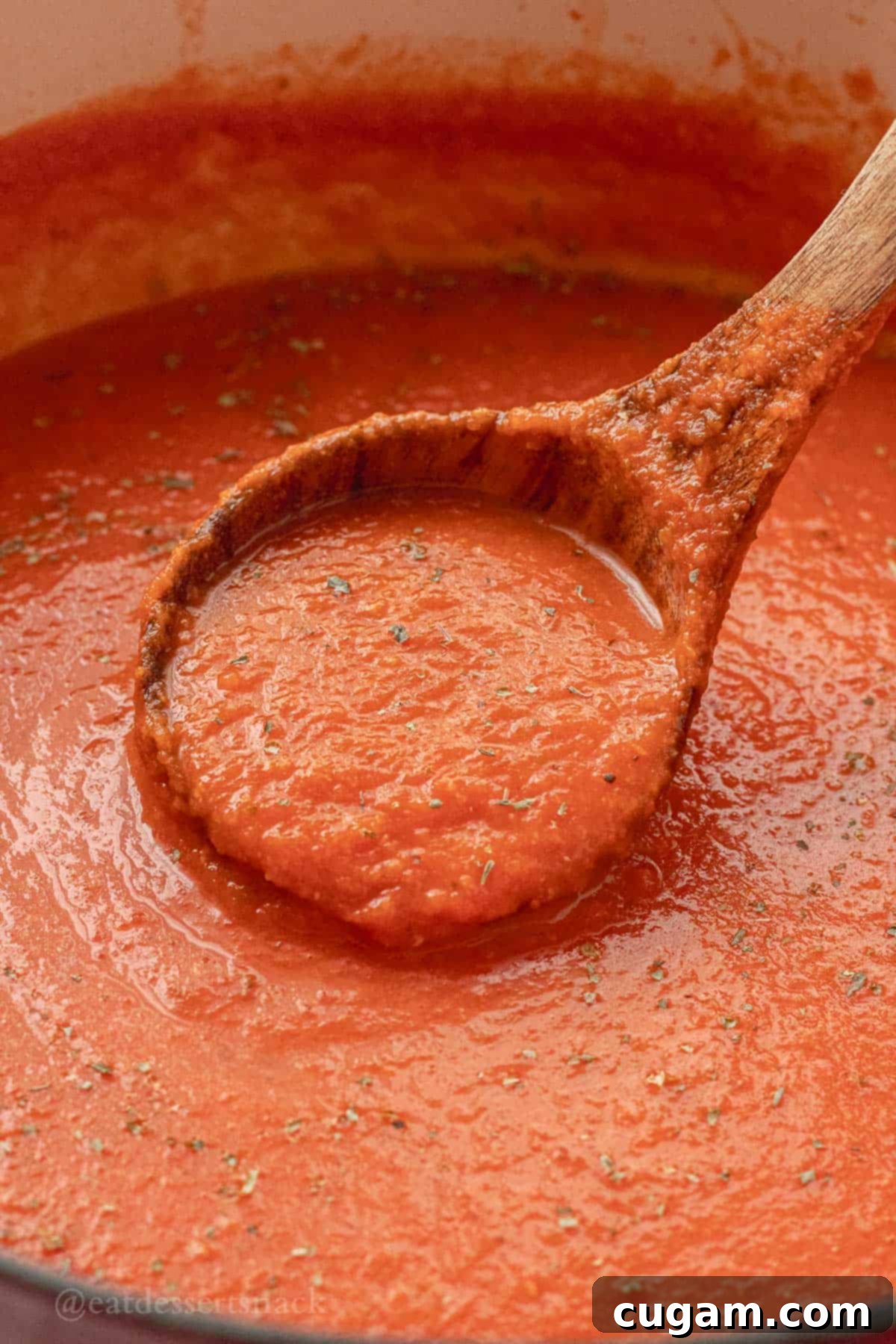 A wooden ladle holding a portion of cooked homemade tomato soup, garnished with fresh basil, over a red pot.