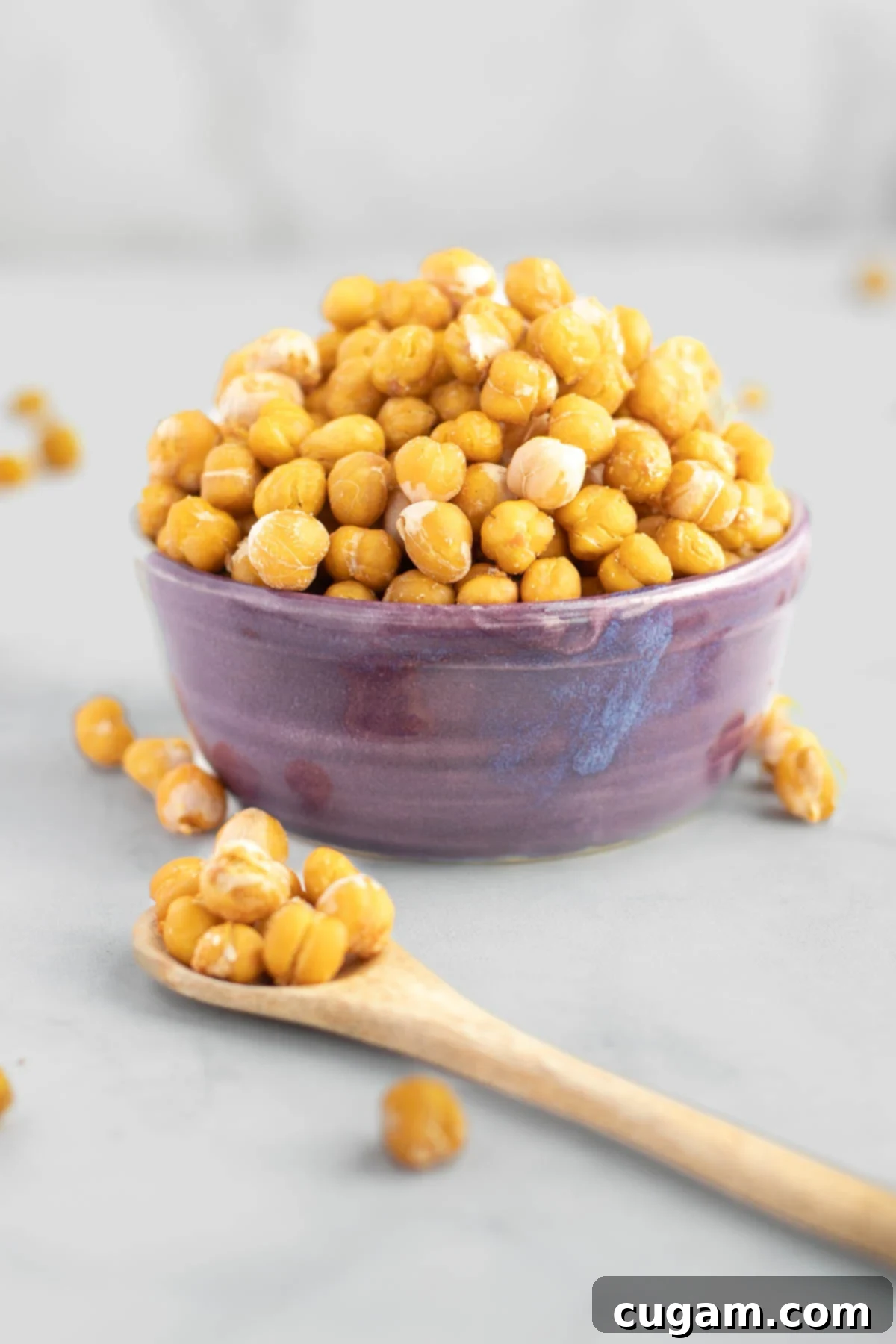 Crunchy roasted chickpeas in a vibrant purple bowl. A spoonful of chickpeas is held close up in front of the bowl.
