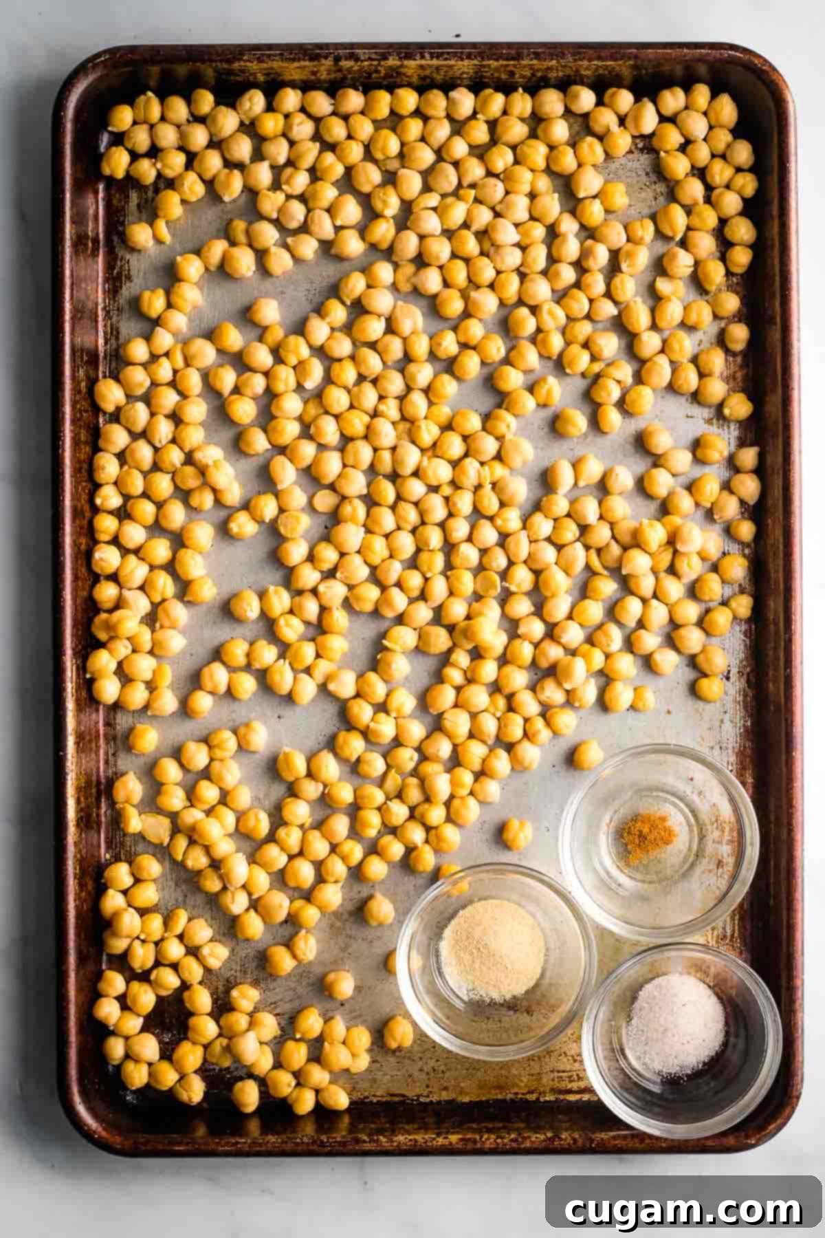 Chickpeas spread in a single layer on a baking sheet, with small dishes of garlic powder, cayenne pepper, and sea salt nearby.