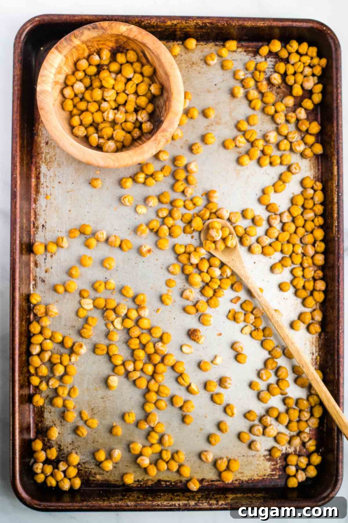 A spoon is scooping crunchy chickpeas from a baking tray into a purple serving bowl.