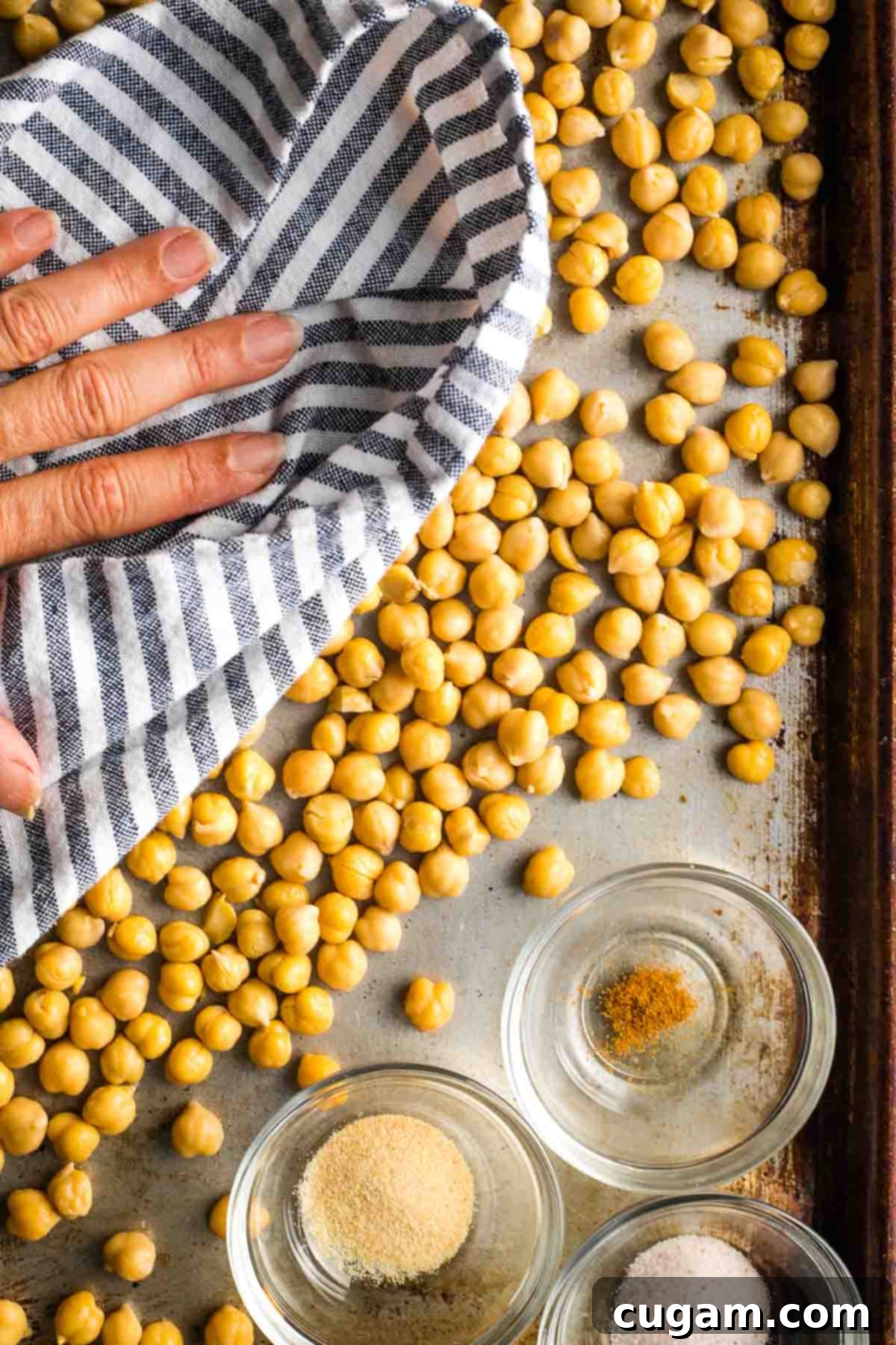 A hand is gently rubbing chickpeas on a baking sheet with a paper towel, emphasizing the drying process.