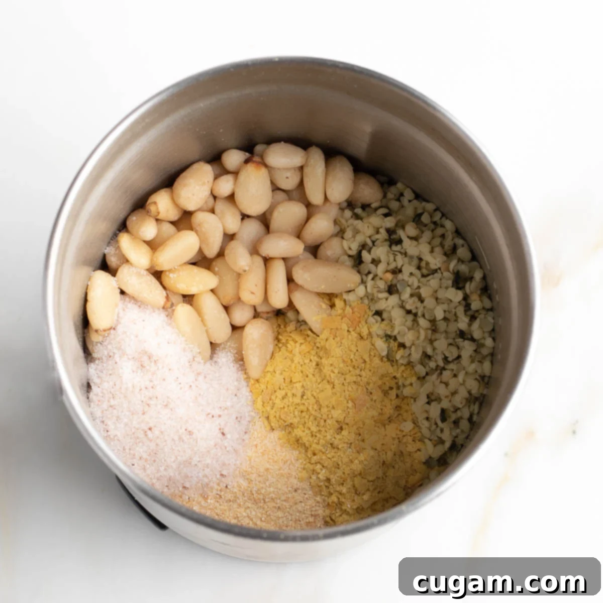 Ingredients in the bowl of a spice grinder: pine nuts, hemp seeds, nutritional yeast, garlic powder and salt.