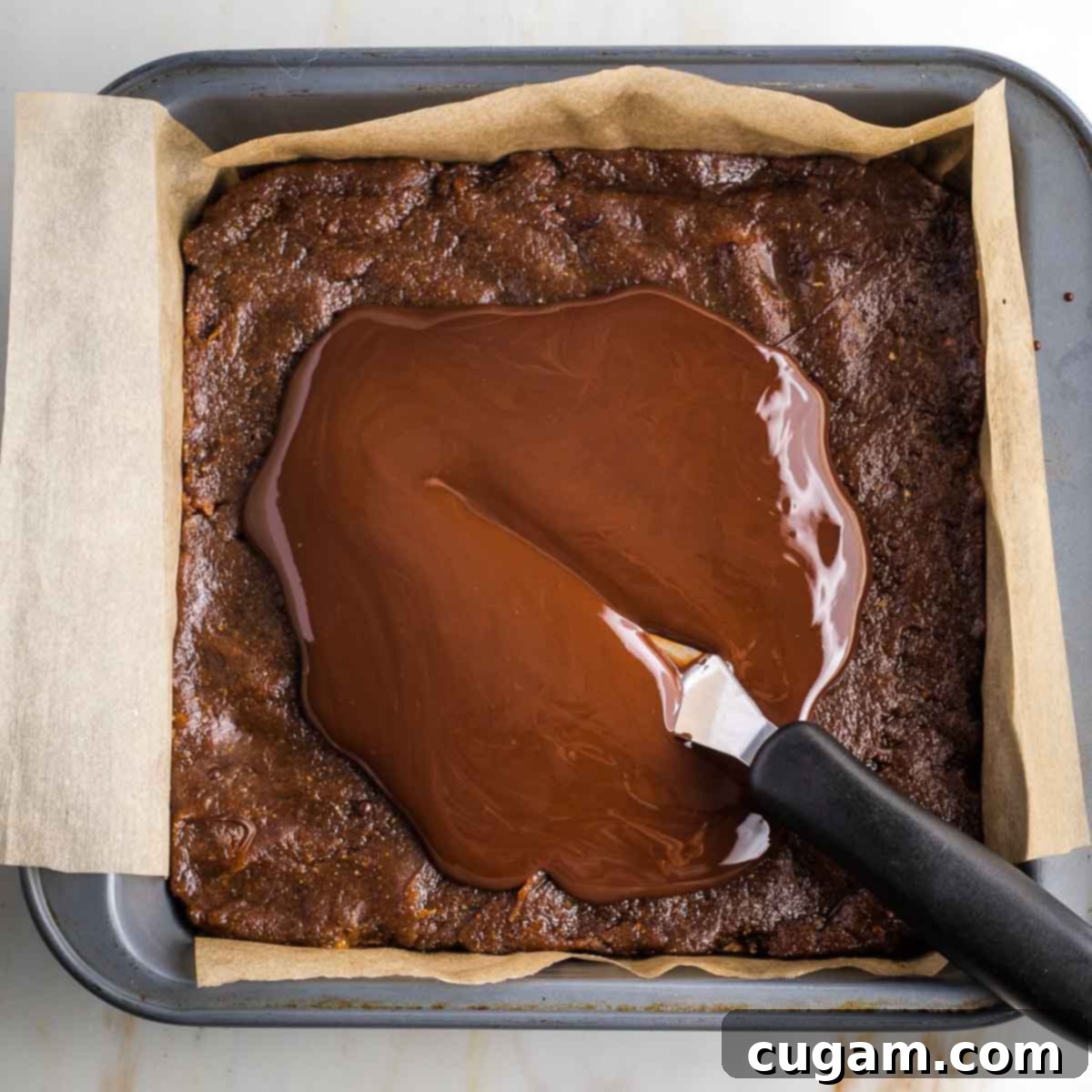 Offset spatula spreading melted chocolate over a caramel layer in a square baking pan.