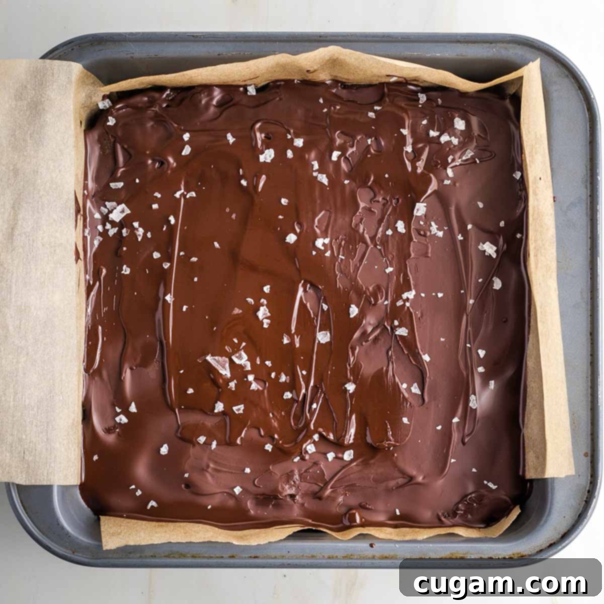 Square baking pan filled with homemade twix, chocolate layer on the top is sprinkled with flakey salt.