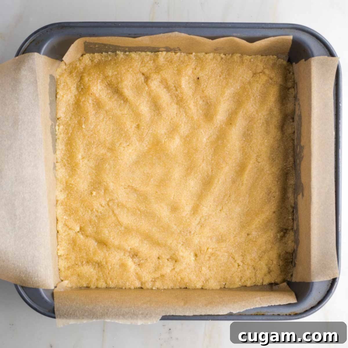 Cookie dough pushed down and into the corners of a square baking pan lined with parchment paper.