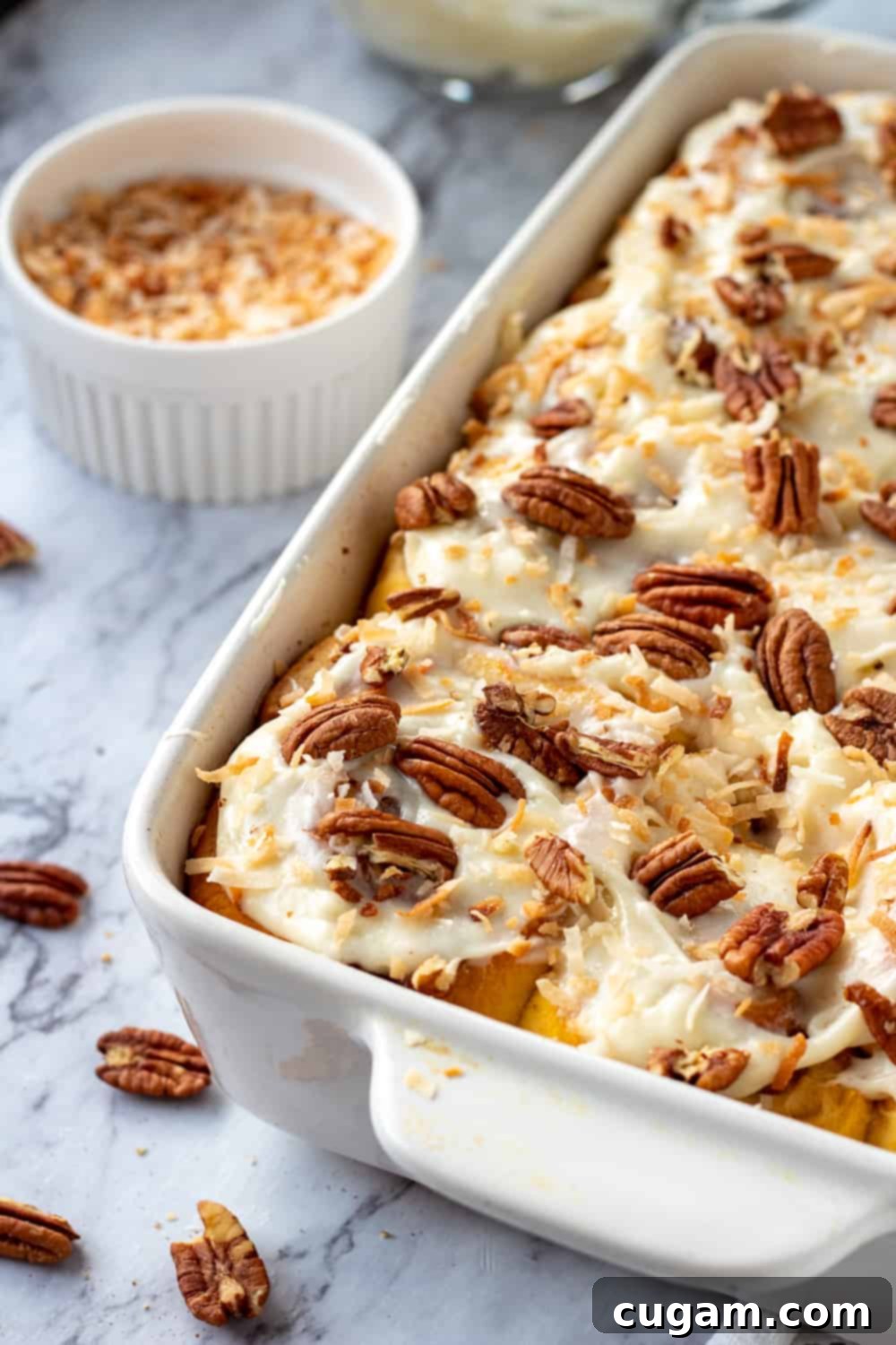 Baked carrot cake cinnamon rolls in white pan with pecans and coconut on marble. 