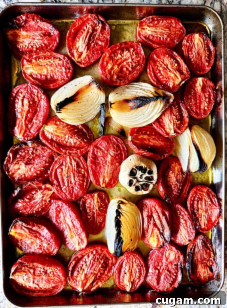 Close-up of Roma tomatoes, quartered Vidalia onion, and a head of garlic on a baking sheet, drizzled with olive oil and ready for roasting.