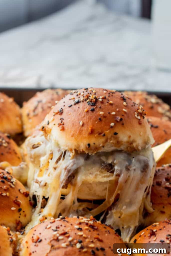 A French Onion Ground Beef Slider, sliced in half, with a perfect cheese pull stretching from the melted cheese and filling, lifted with a gold fork. 
