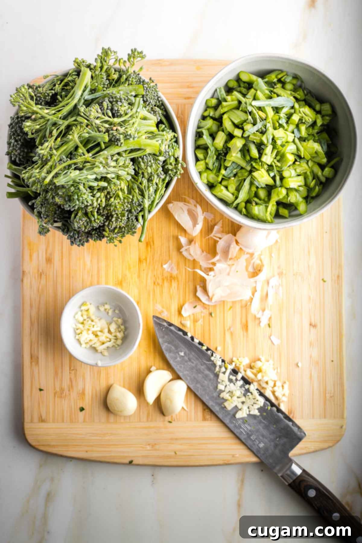 Knife mincing garlic on a board that has bowls of trimmed broccolini.