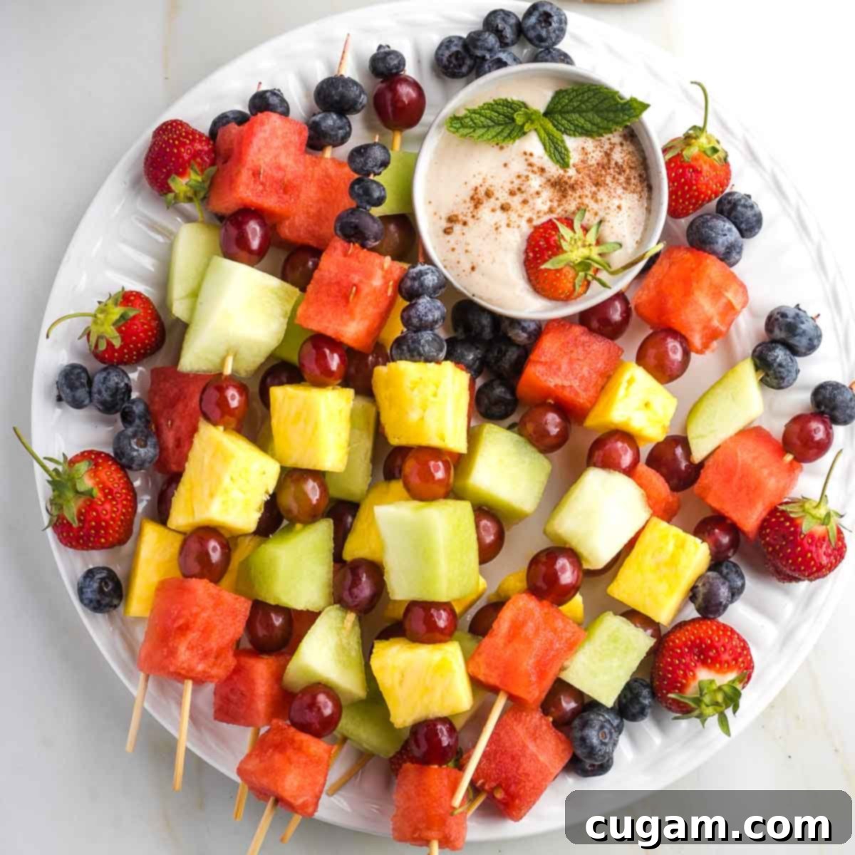Skewers of colorful fruit with a small bowl of creamy dip, sprinkled with cinnamon. 