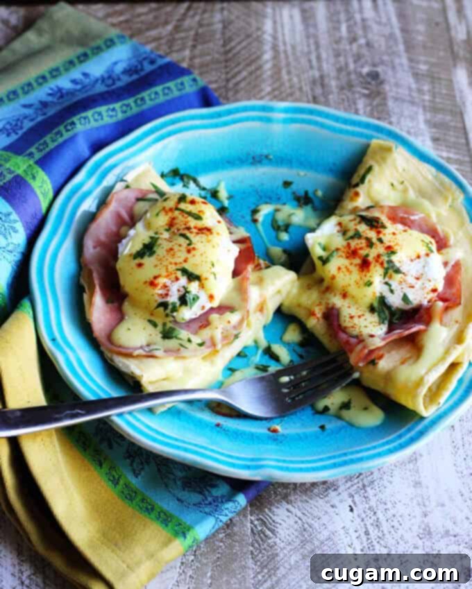 A close-up of a stack of Crepes Eggs Benedict with runny yolk and Hollandaise sauce.